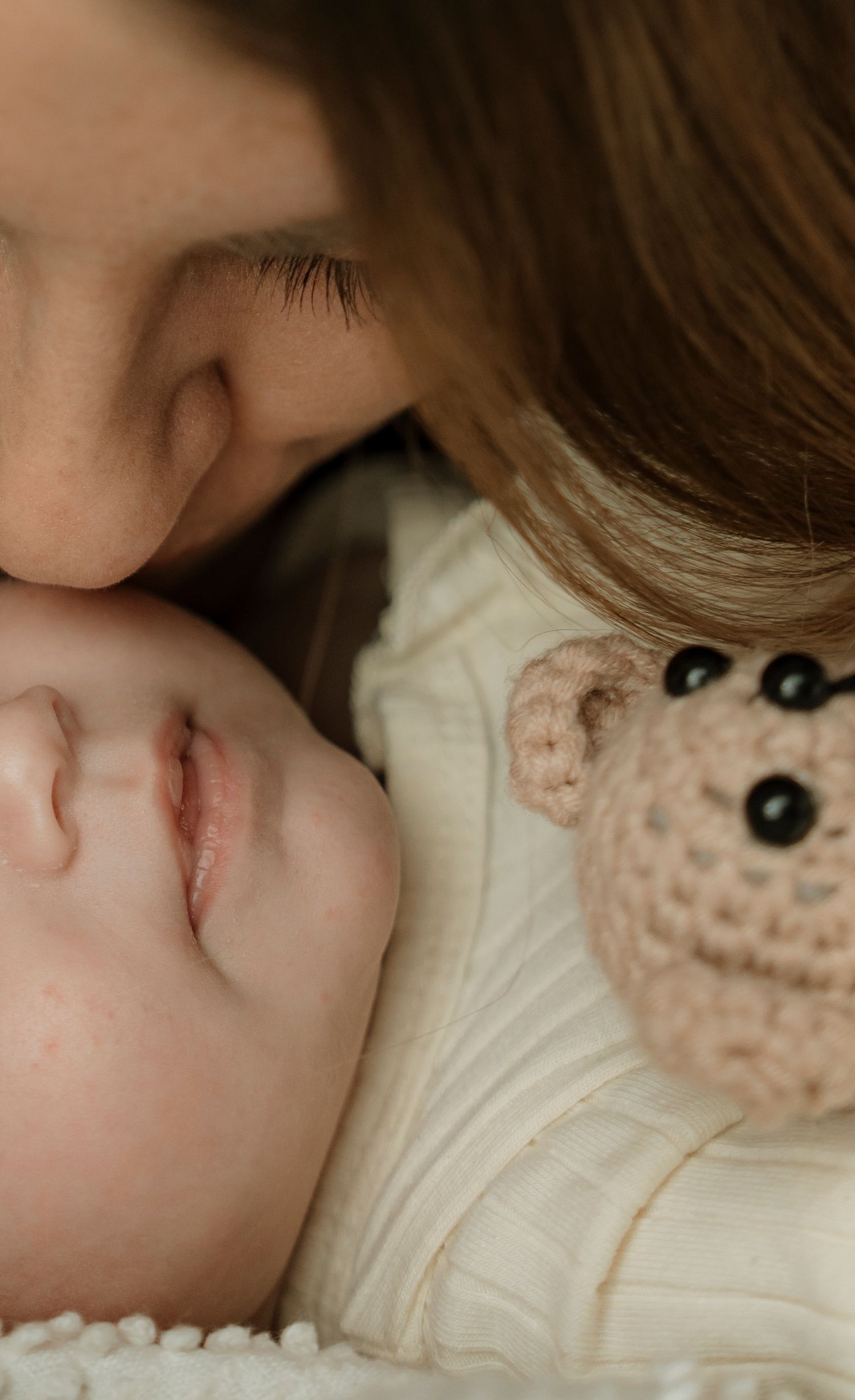 A tender moment as a mother gently kisses her sleeping newborn baby beside a cute crocheted bear toy.