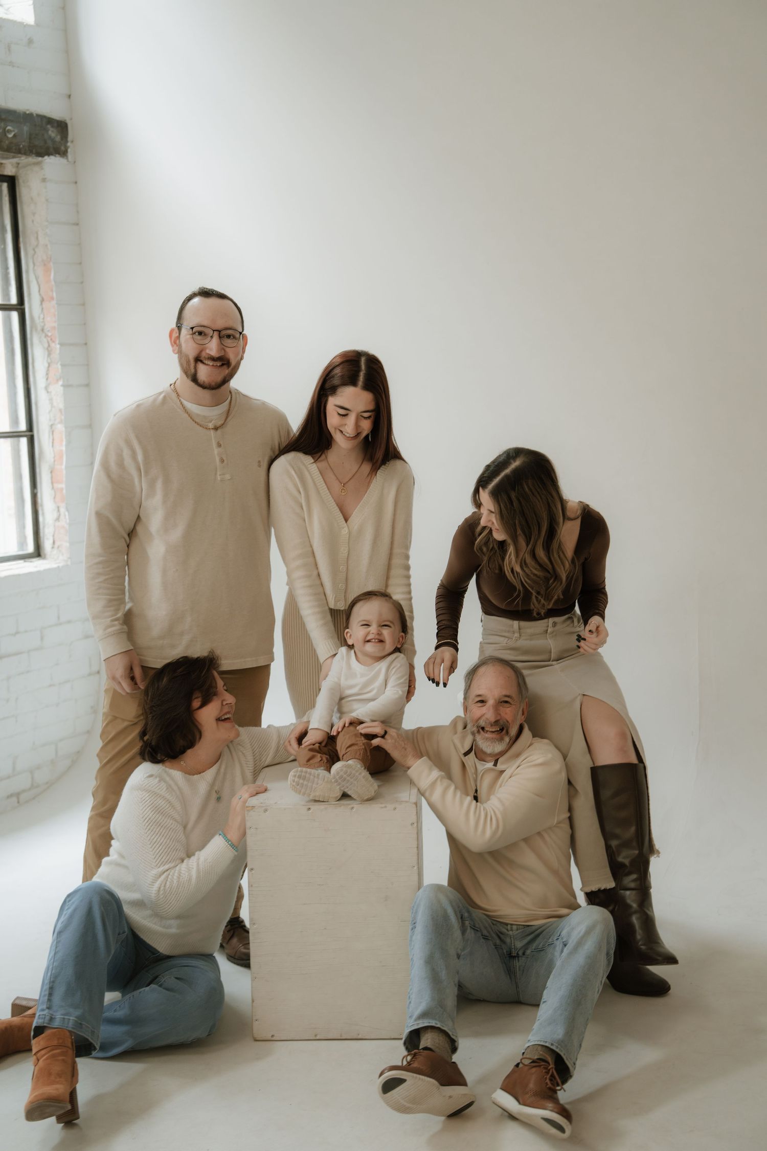 A family of three poses in a bright studio, with parents crouching beside their baby in matching neutral tones.