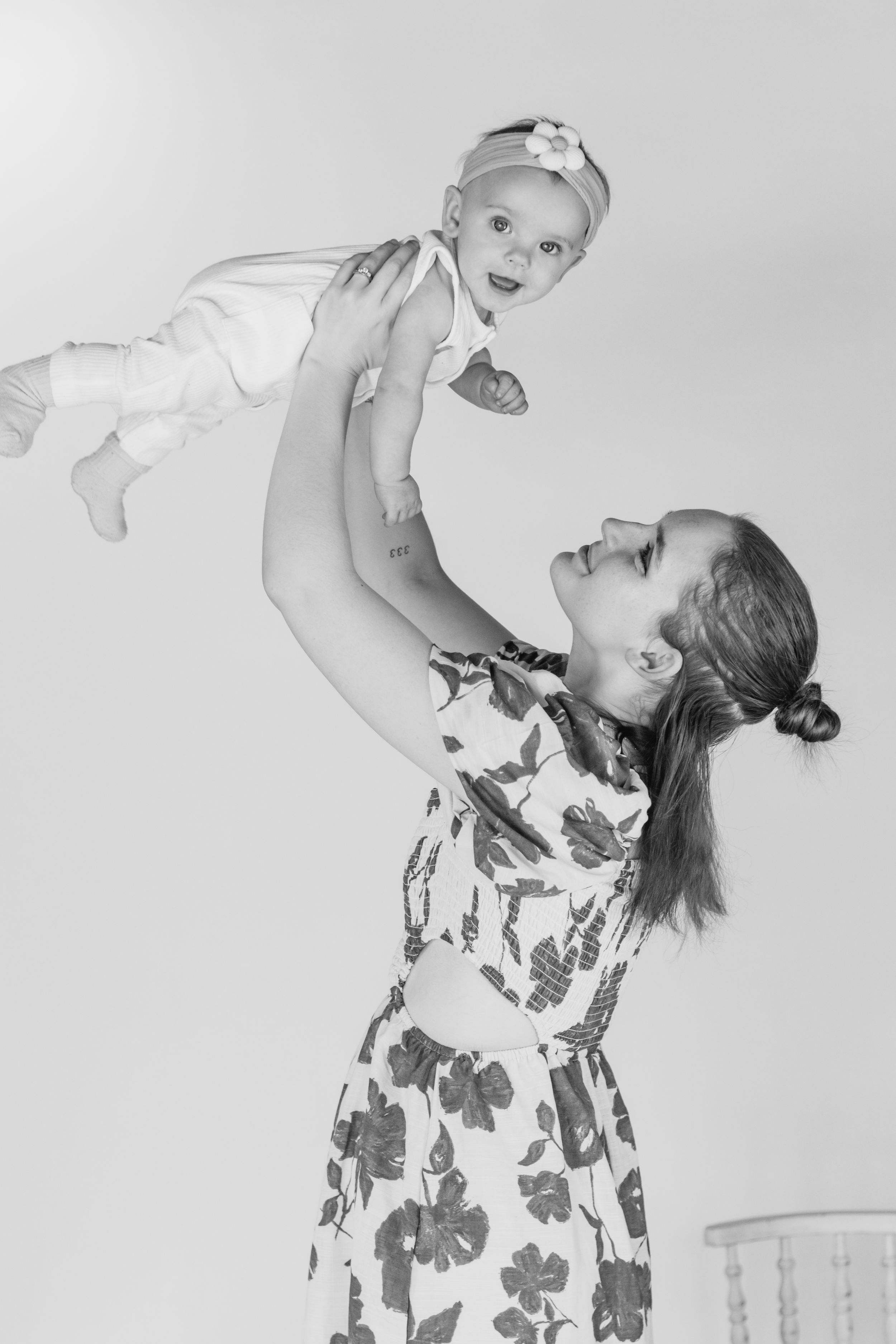 Black and white photo of mother lifting laughing baby girl overhead in floral dress captured by pierce county photographer