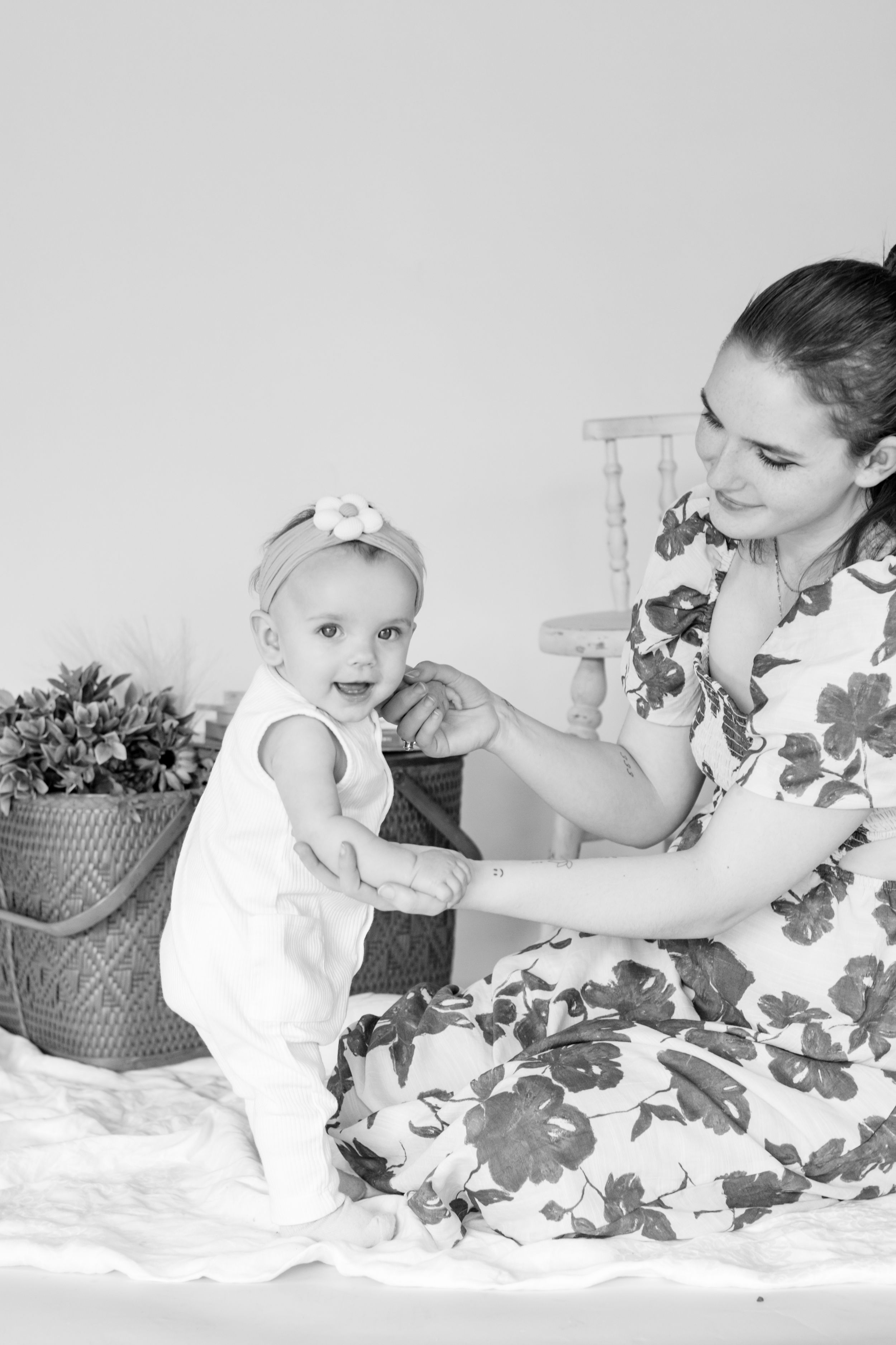 Black and white photo of mother helping baby girl stand while sitting on white blanket at indoor studio in roy wa