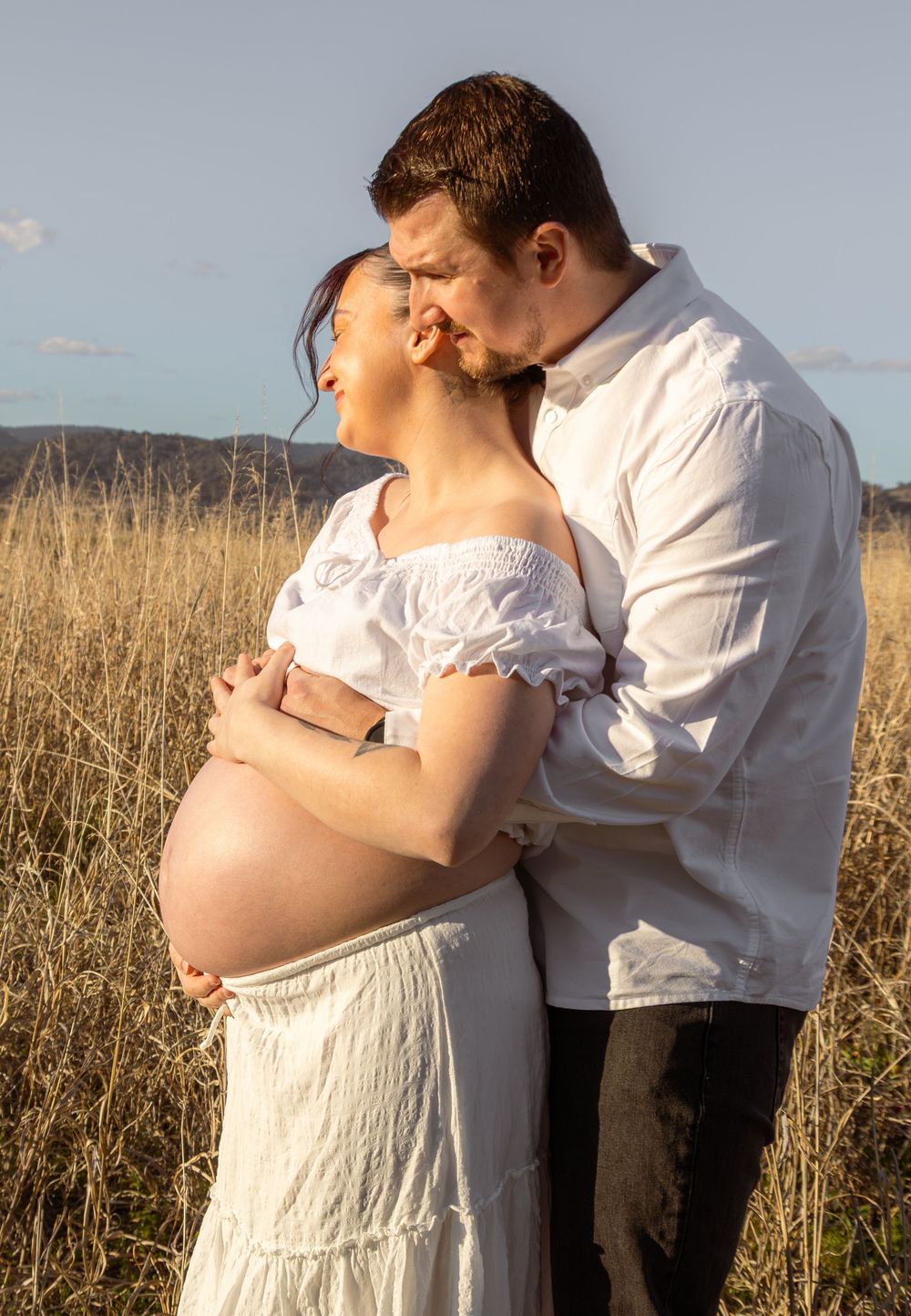 Pregnant couple embracing in golden field, woman in white off-shoulder dress, man kissing her cheek at sunset.