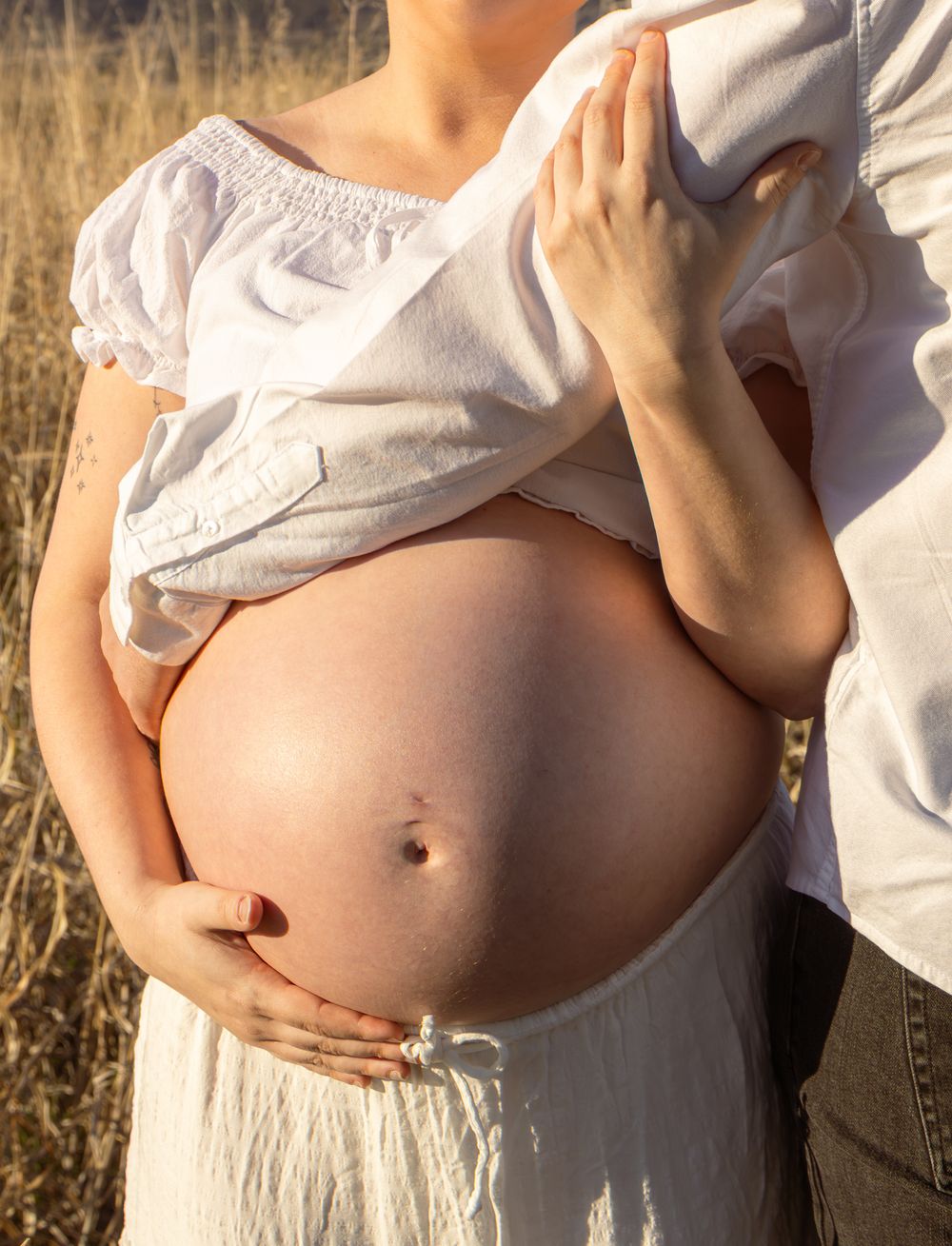 Pregnant woman in white dress outdoors with partner cradling her bare baby bump in a sunny field.