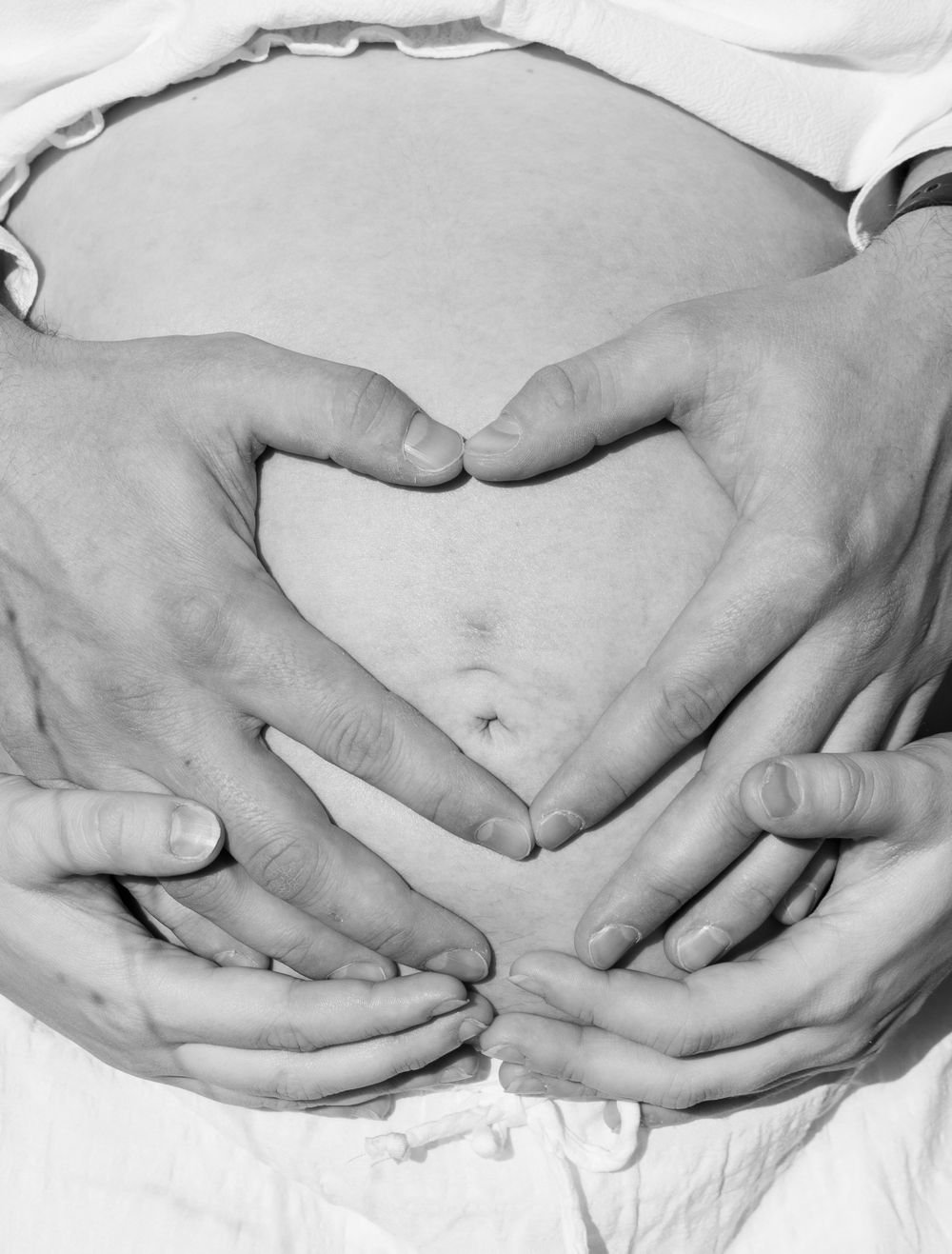 Two pairs of hands forming a heart shape on a pregnant belly in a black and white maternity photo.
