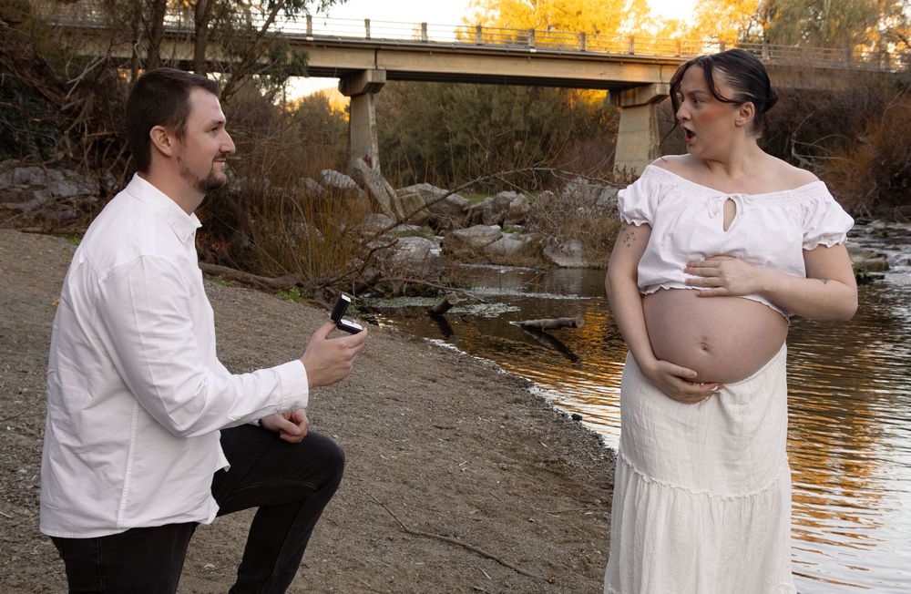 Pregnant woman in white dress with man on one knee proposing by a river with a bridge in the background.