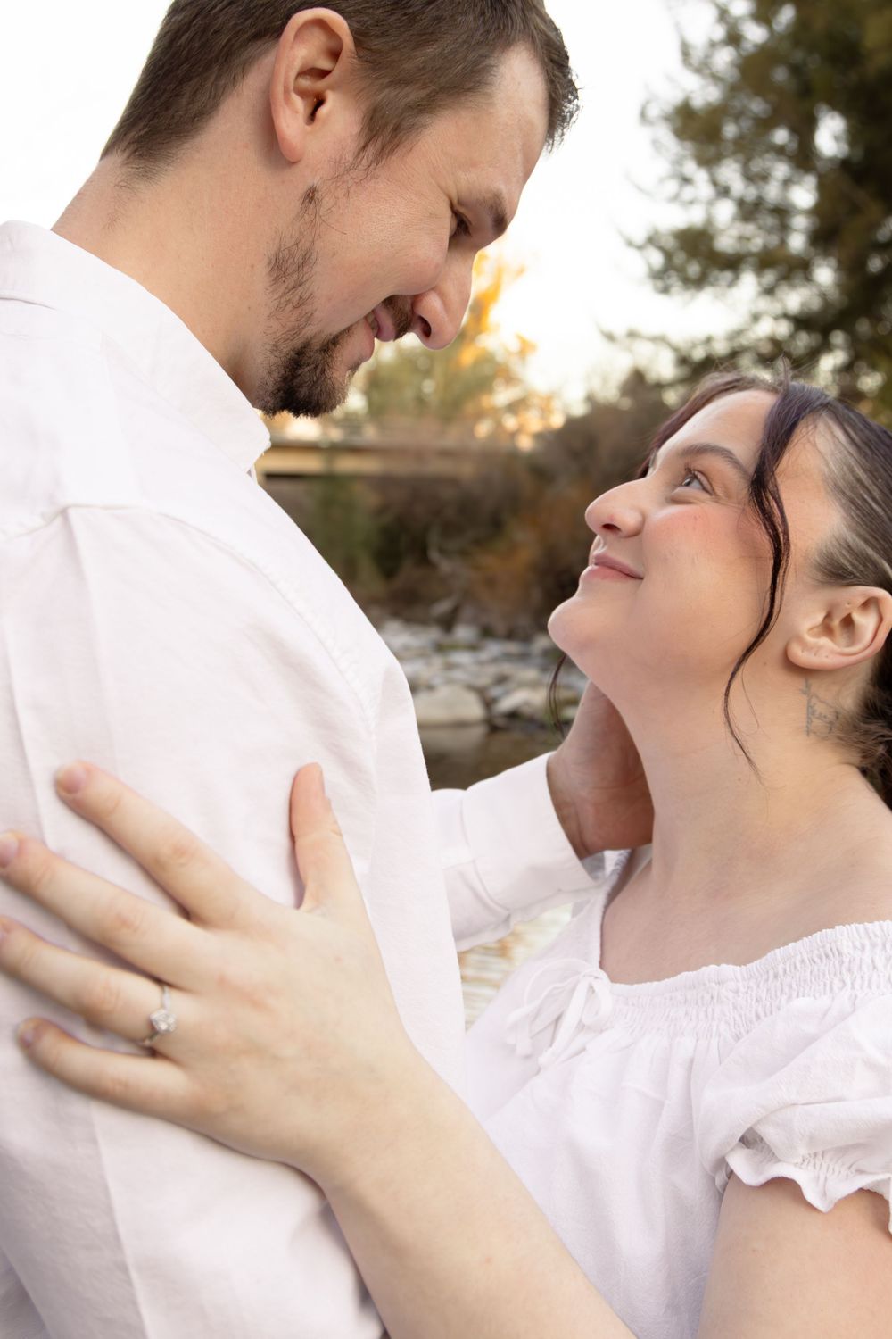 Couple in white outfits gazing at each other outdoors, woman wearing engagement ring, romantic portrait.
