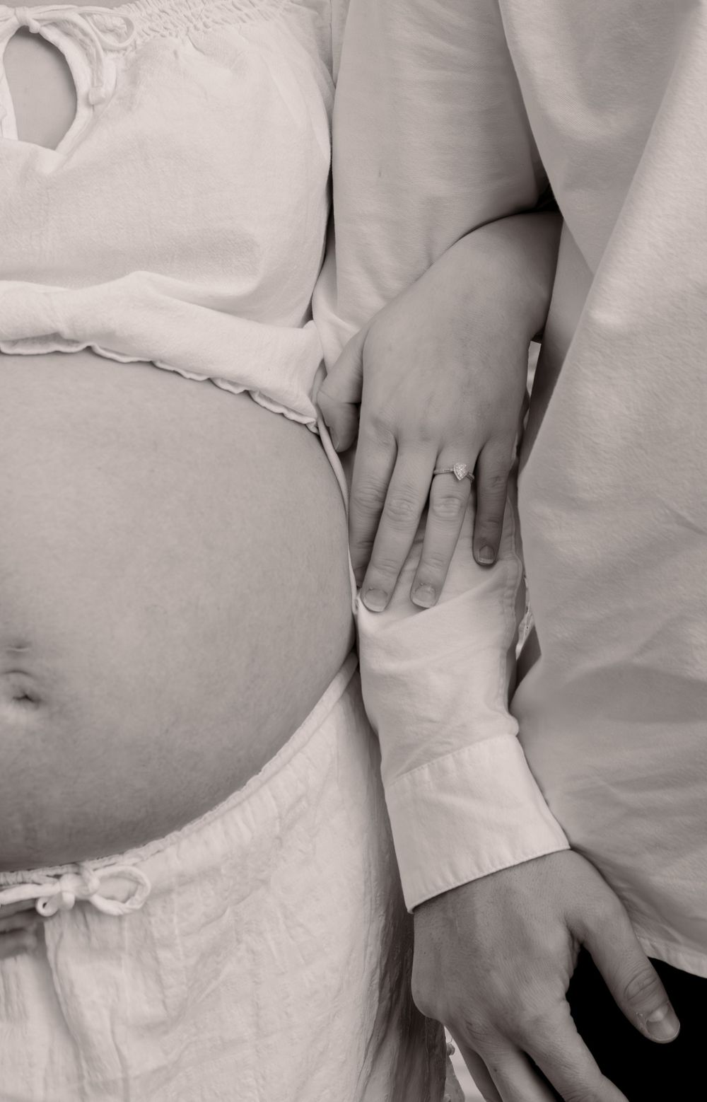 Black and white photo of pregnant woman with partner's hand resting on her baby bump, wearing white clothing.
