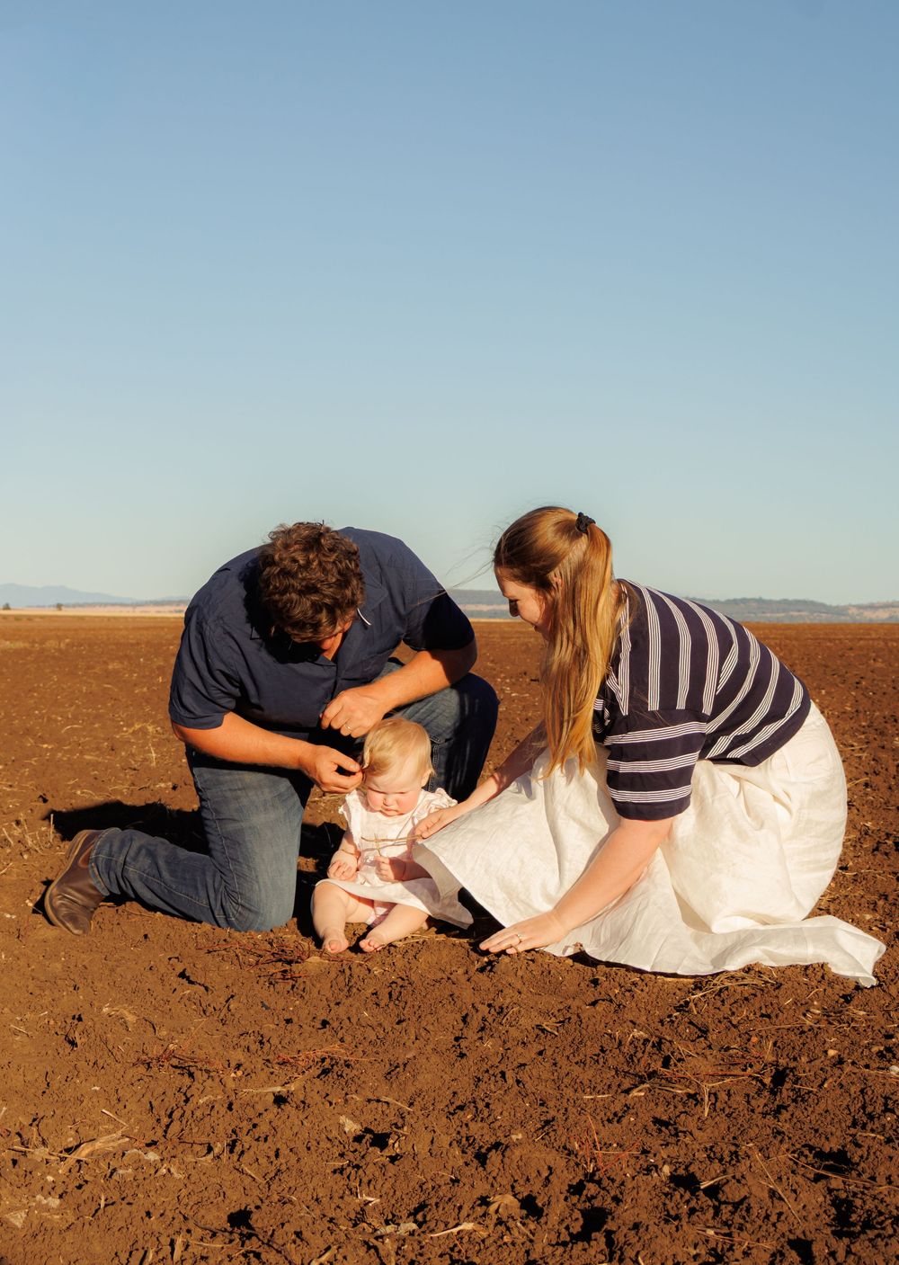 Parents kneeling in red dirt field helping their baby sit on the ground.