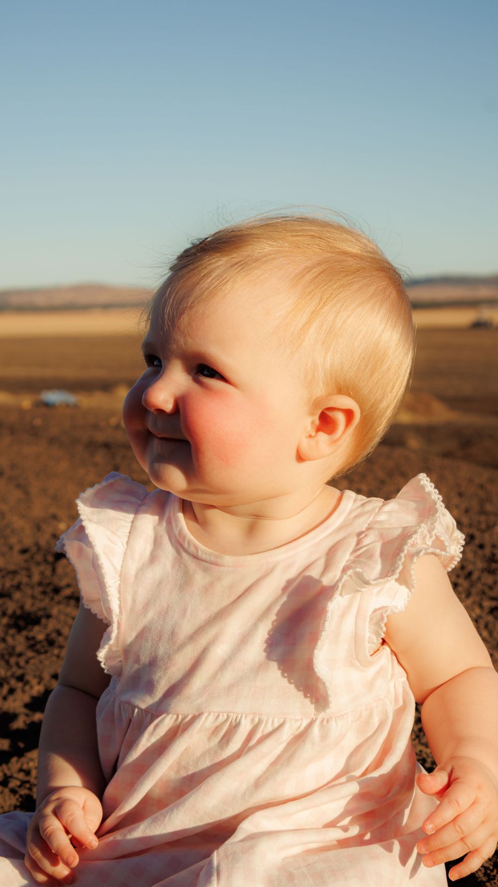 Smiling blonde baby girl in white dress sitting outdoors in a farm field.