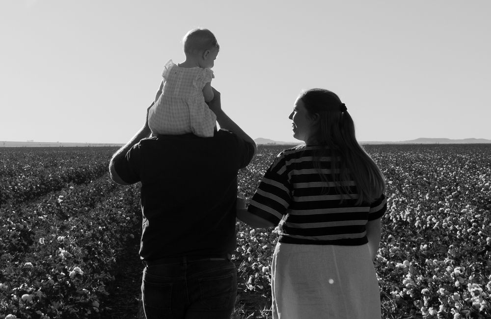 Black and white photo of parents holding baby on shoulders in cotton field.