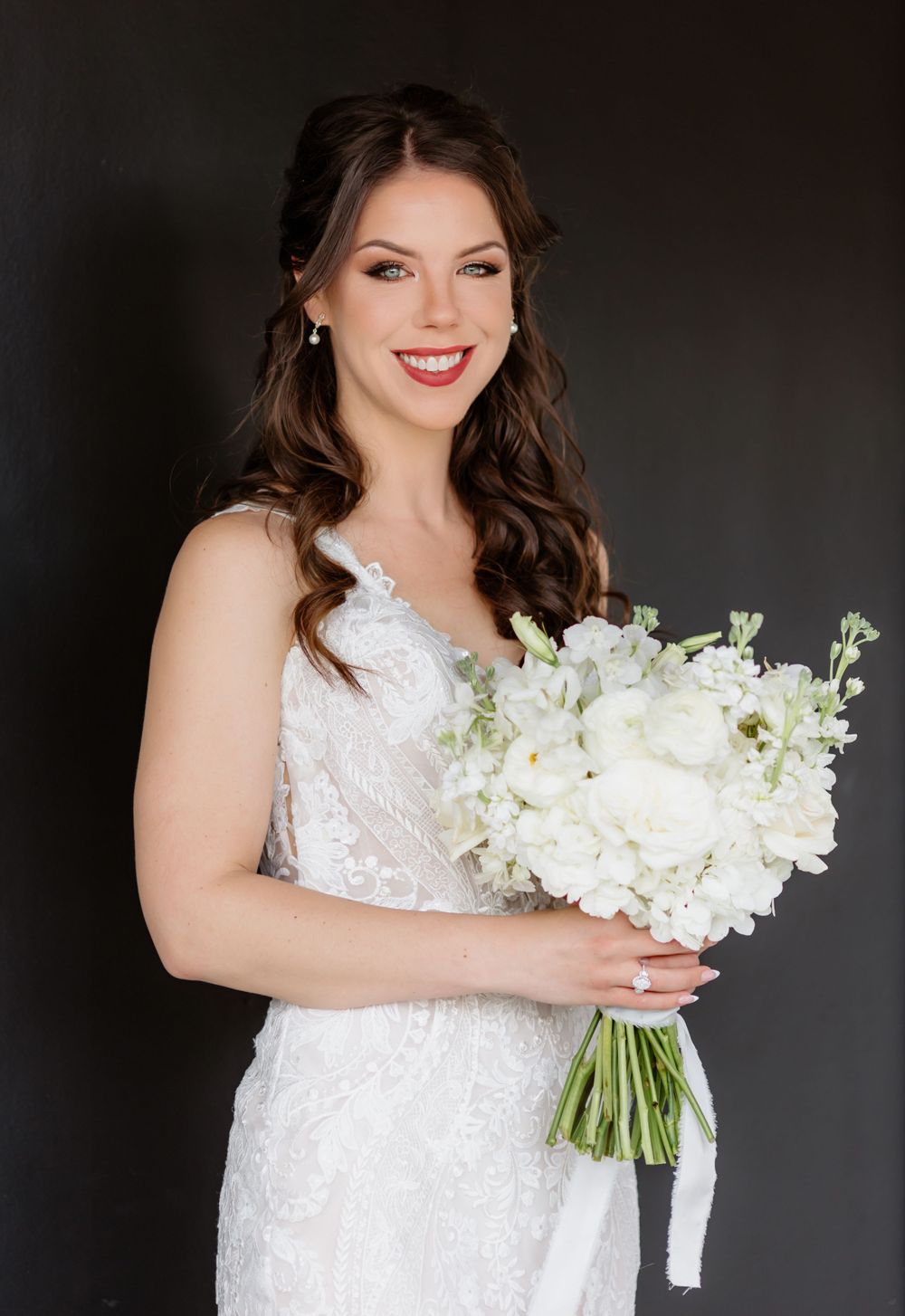 Smiling bride in lace wedding gown holding white floral bouquet against dark background.