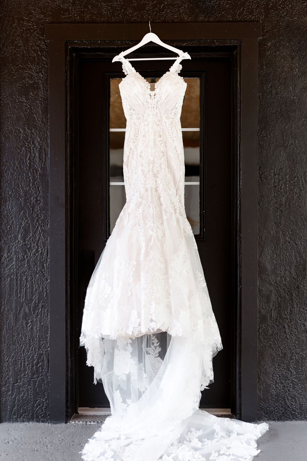 Elegant lace mermaid wedding dress with train hanging on a black door frame.