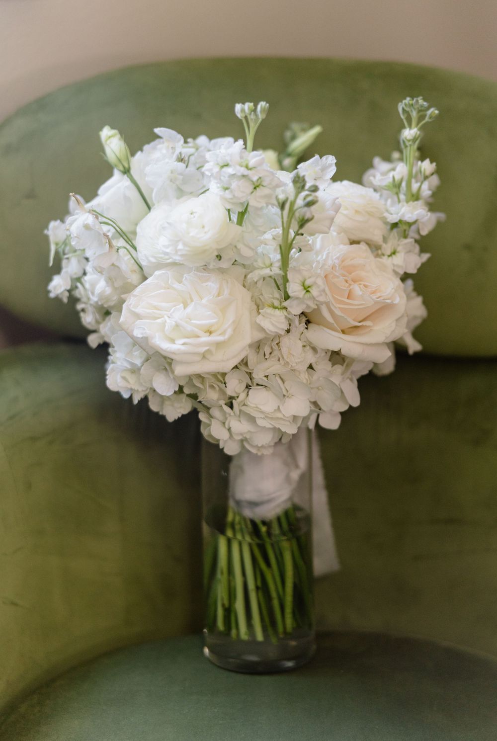 Elegant white and blush bridal bouquet with roses and stock flowers in a glass vase on a green velvet chair.