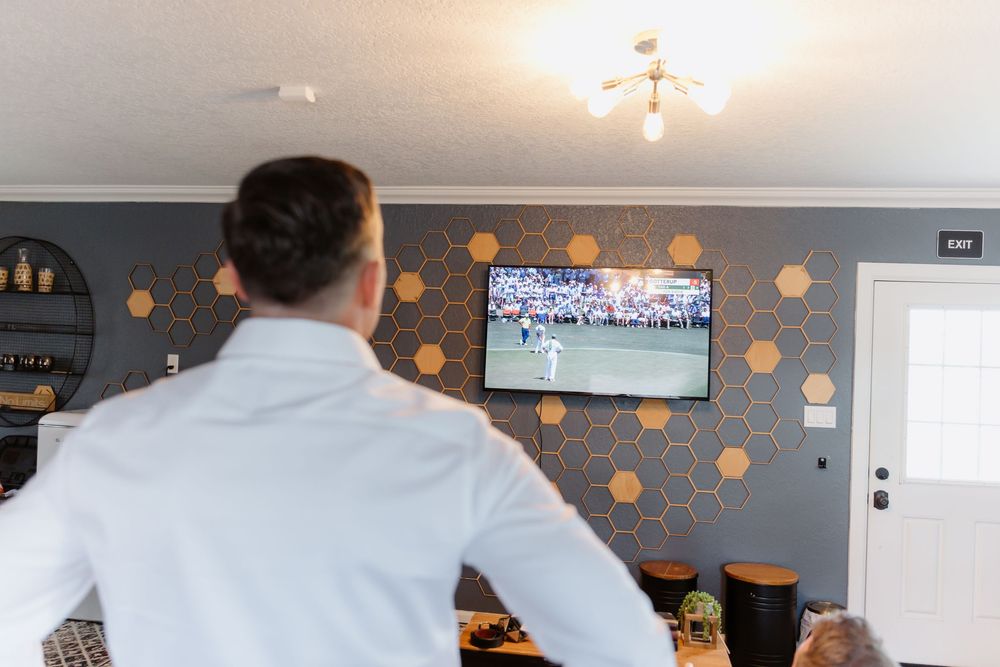 Man in white shirt watching sports game on wall-mounted TV in modern bar with hexagonal pattern wall decor.