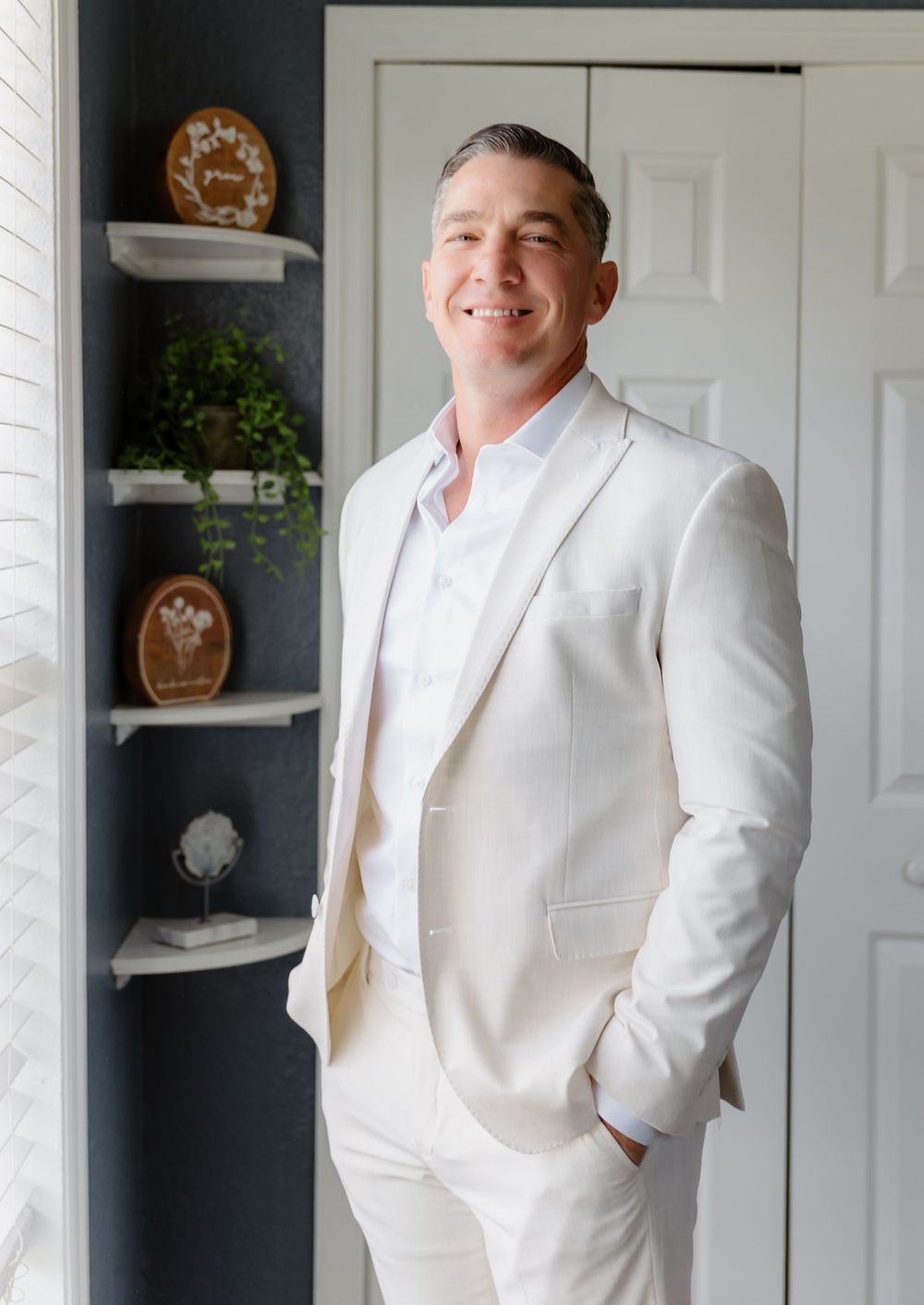 Man in white suit smiling confidently indoors near decorative shelves with greenery and wooden accents.