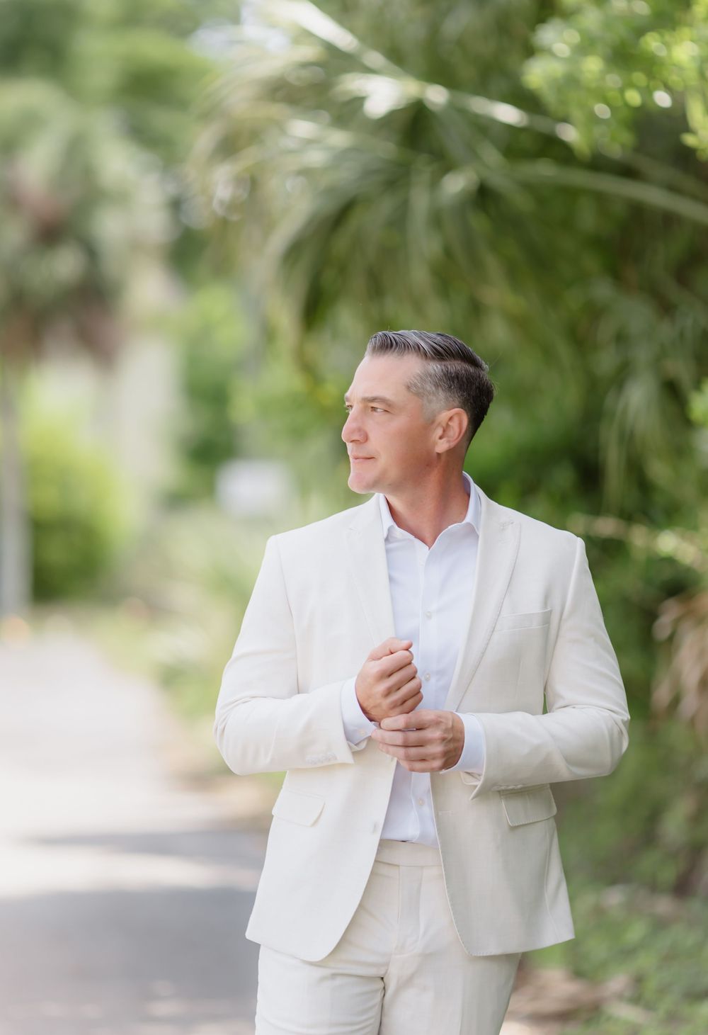 A well-dressed man in a white suit walks along a tree-lined path, looking confidently to the side.