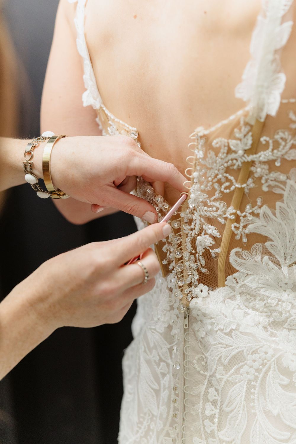 Hands fastening delicate pearl buttons on the back of an intricate lace wedding dress.