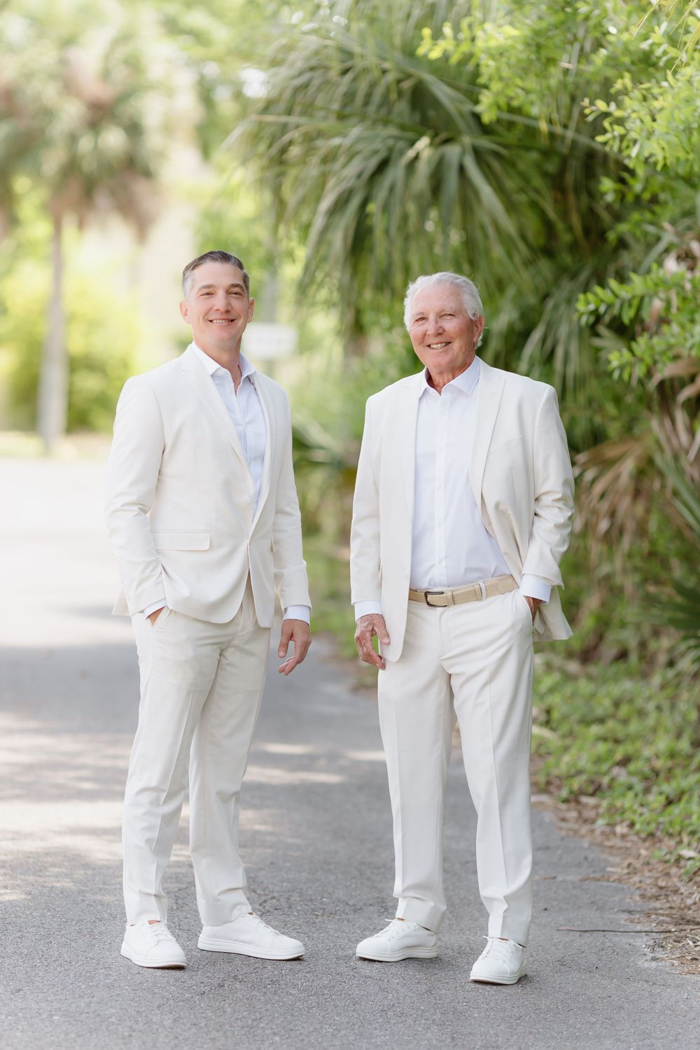 Two men in matching ivory suits pose together outdoors on a path surrounded by lush green tropical foliage.
