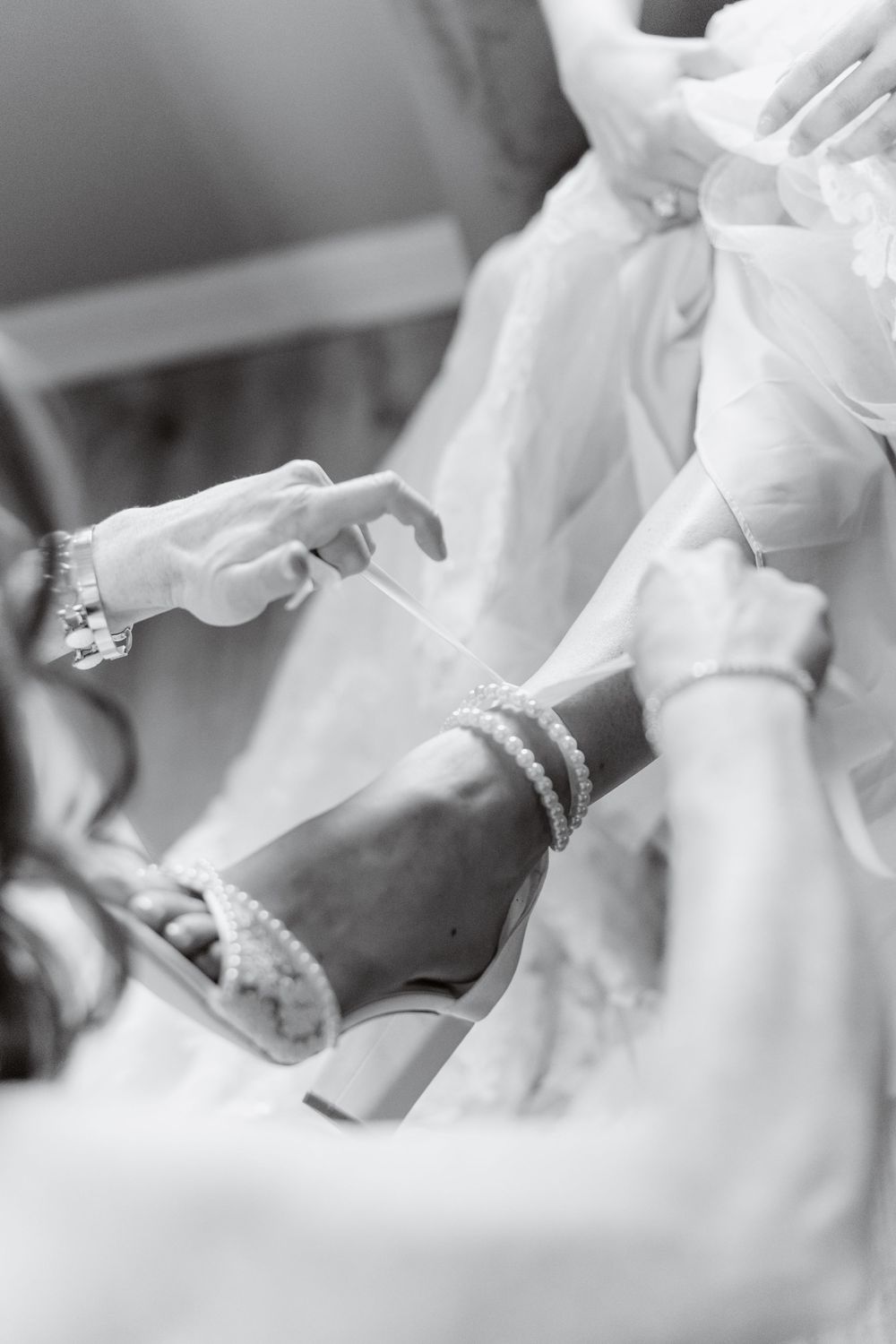 Black and white photo of bride getting pearl ankle strap heels fastened while wearing a white wedding gown.