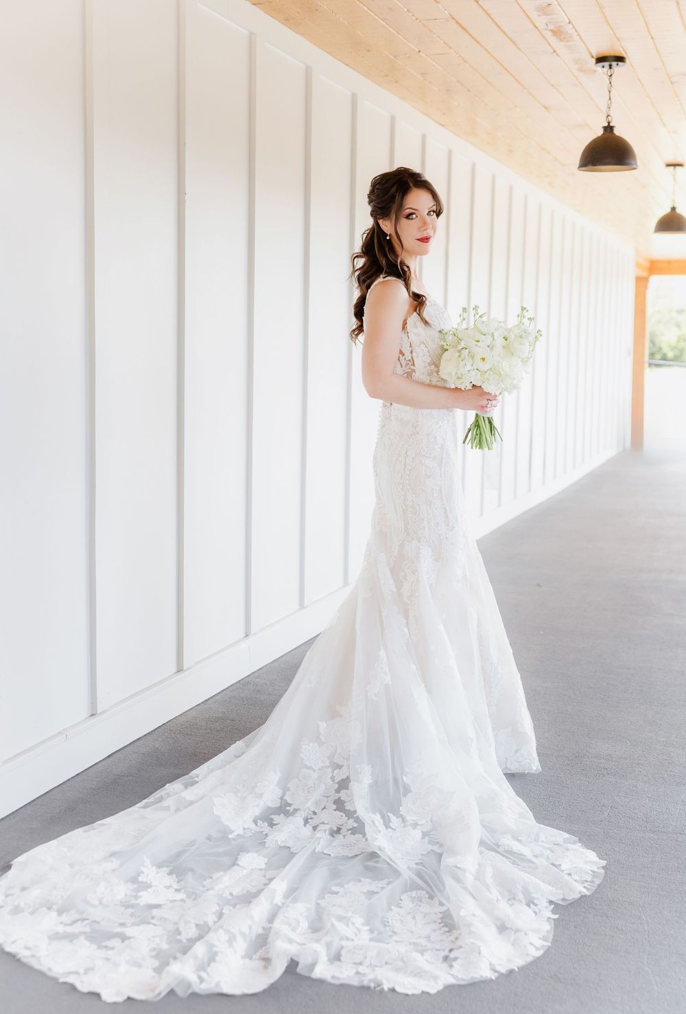 Elegant bride in a lace mermaid wedding gown with long train holds white bouquet in a bright white hallway.