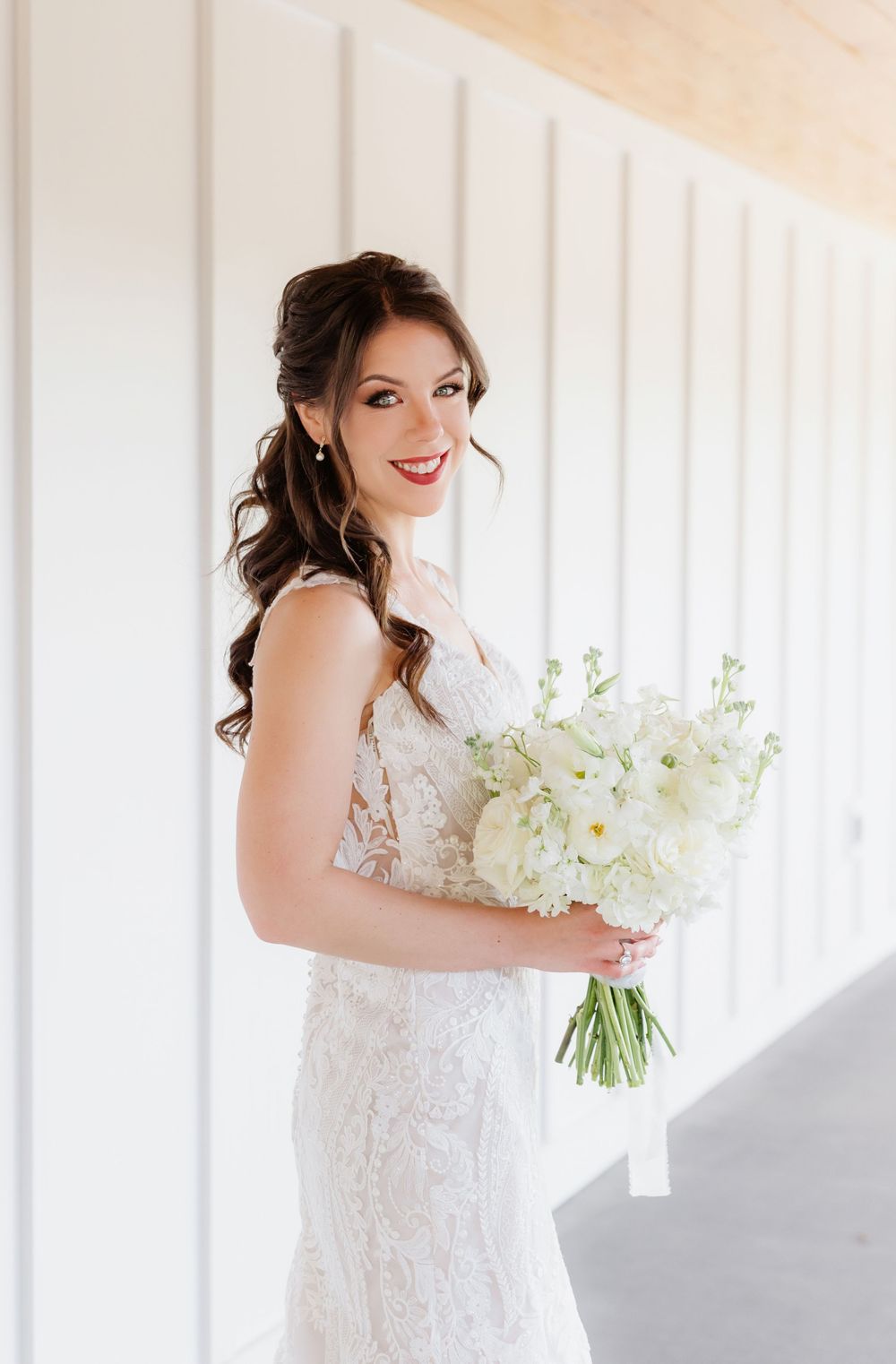 Bride in lace wedding gown holding white floral bouquet, smiling with red lips and elegant half-up hairstyle.