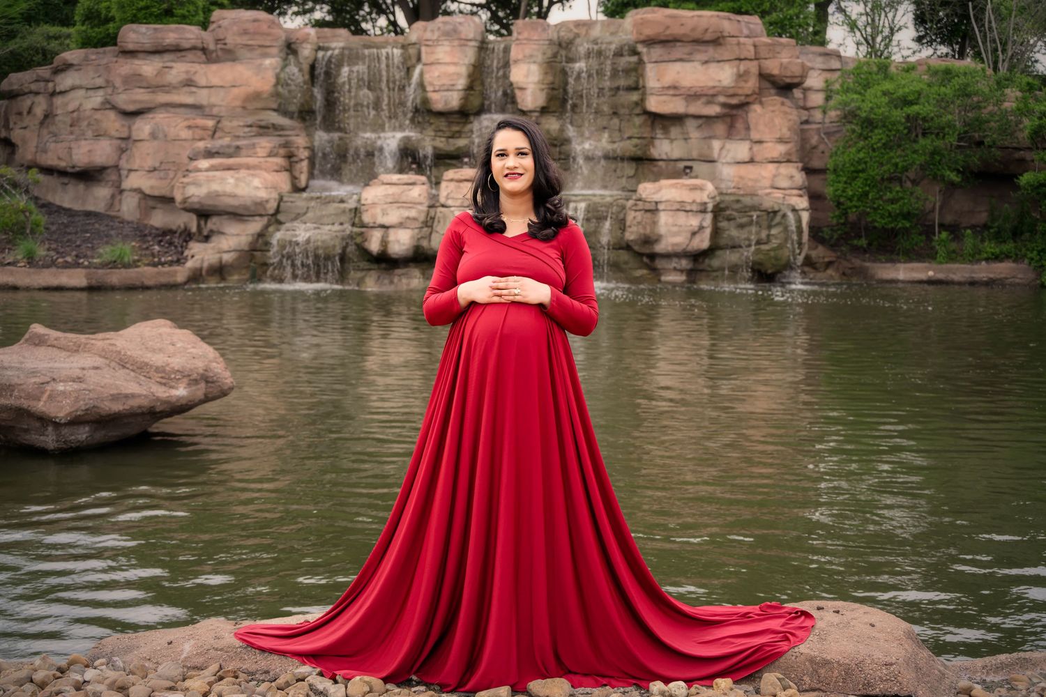Pregnant woman in red maternity gown poses on rocks by waterfall for outdoor maternity photo session. Maternity Photographer in Cypress TX
