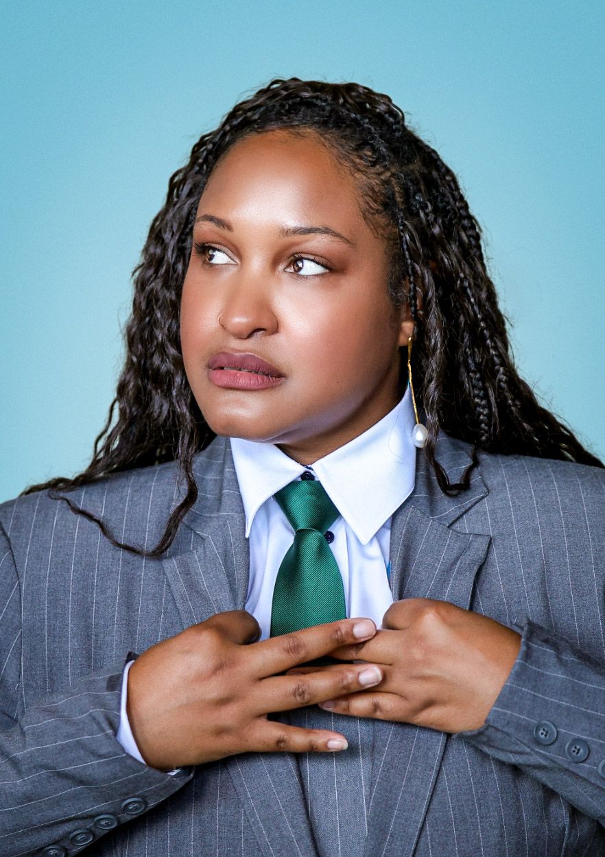 Woman in pinstripe suit and green tie posing against blue background with hands on chest.