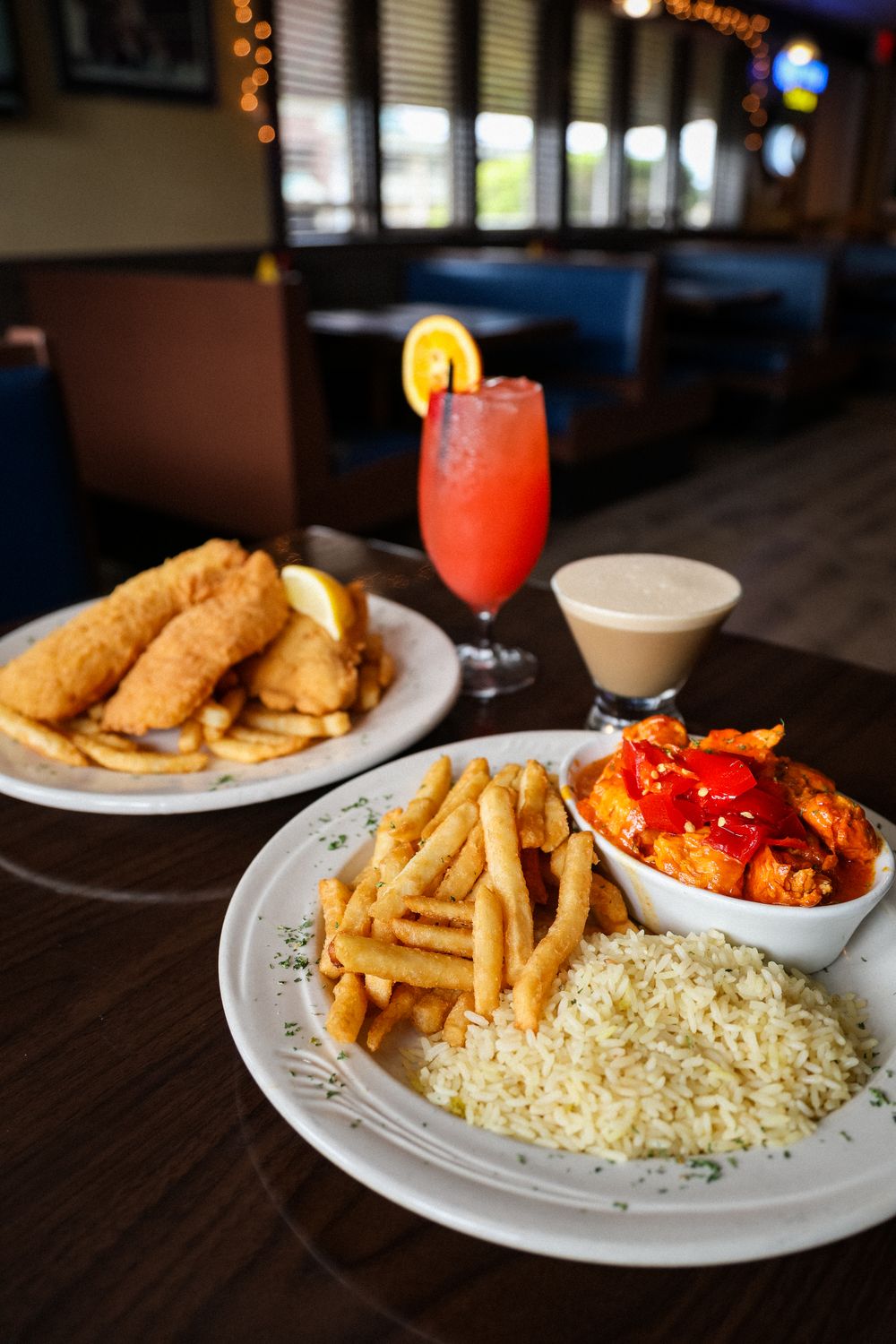 Restaurant table with fried fish, fries, rice, buffalo chicken, a creamy dip, and a red cocktail garnished with orange.
