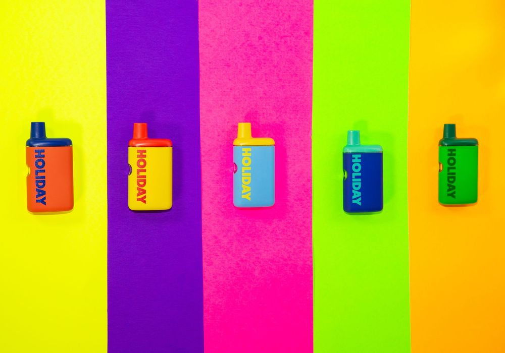 Five colorful Holiday disposable vapes displayed on vibrant striped background in rainbow colors.
