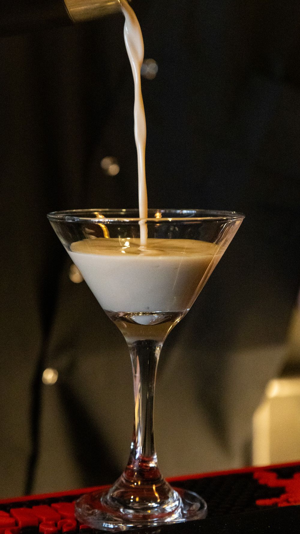 Creamy white cocktail being poured into a martini glass on a dark background.