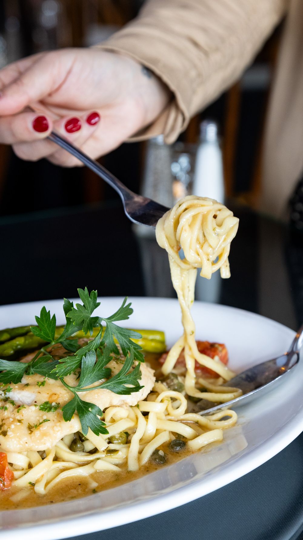 A woman with red nails twirls creamy fettuccine pasta with shrimp, asparagus, and fresh herbs on a fork over a white bowl.