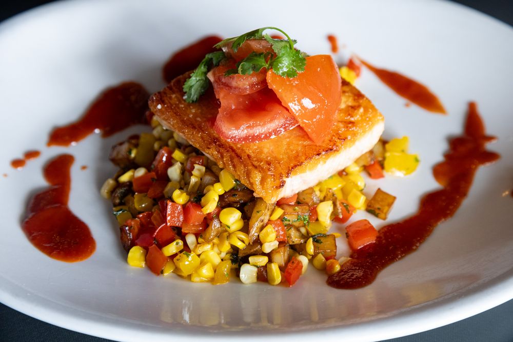 Seared fish fillet on corn succotash with red pepper sauce and fresh tomato garnish on white plate.