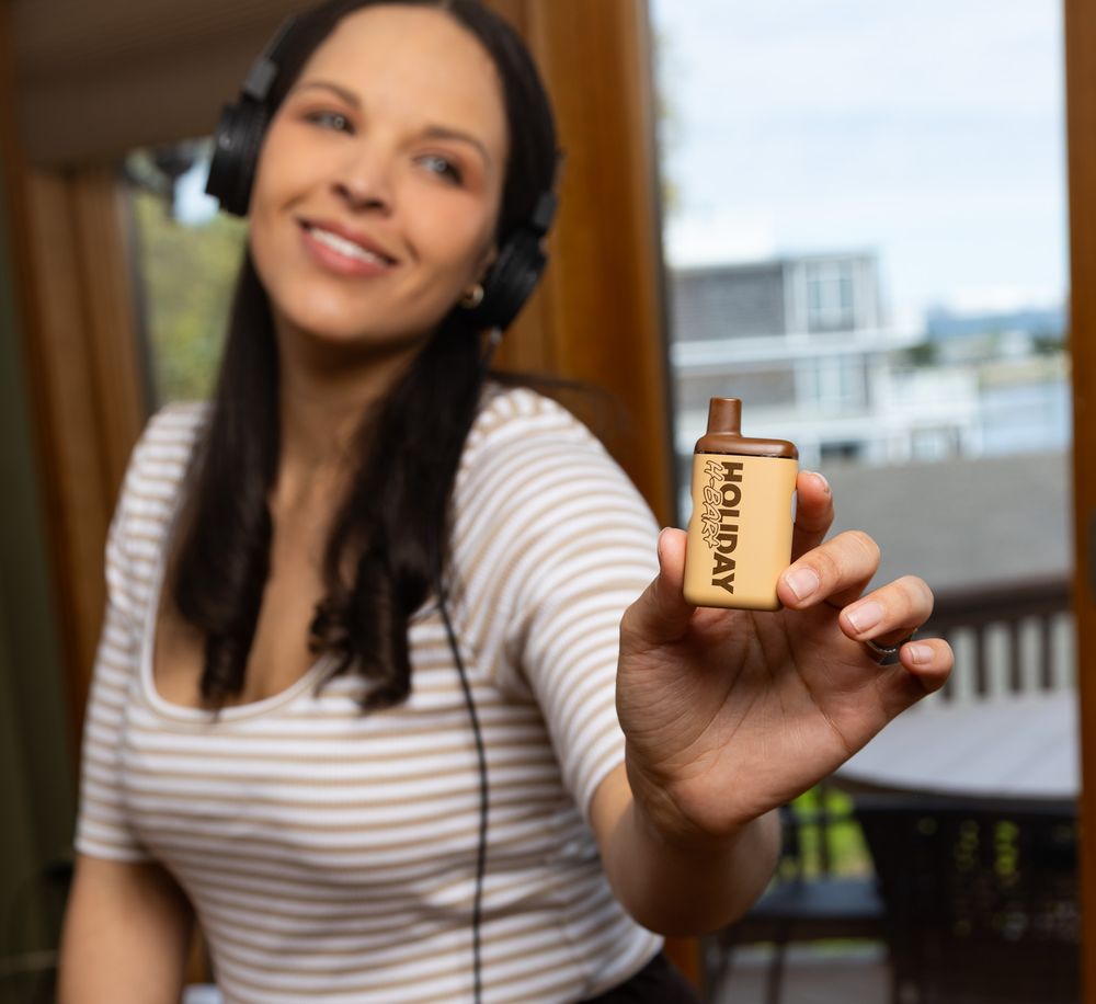 Smiling woman wearing headphones holds up a small Holiday branded bottle outdoors on a deck.
