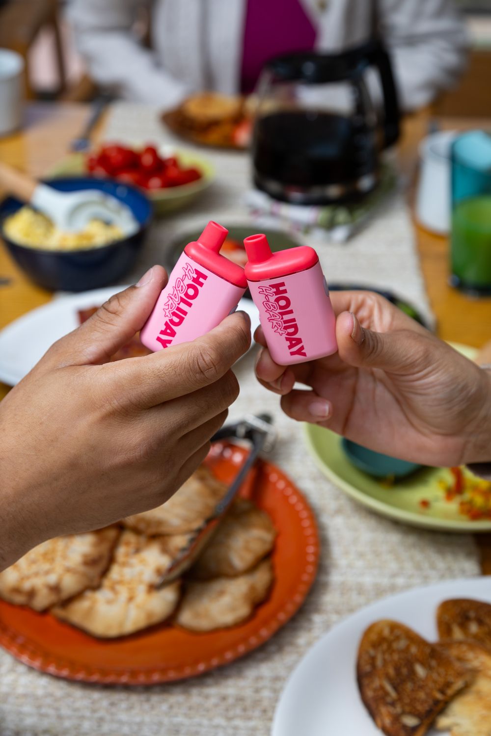 Two hands toasting with pink Holiday branded mini bottles over a breakfast table with toast, fruit, and eggs.