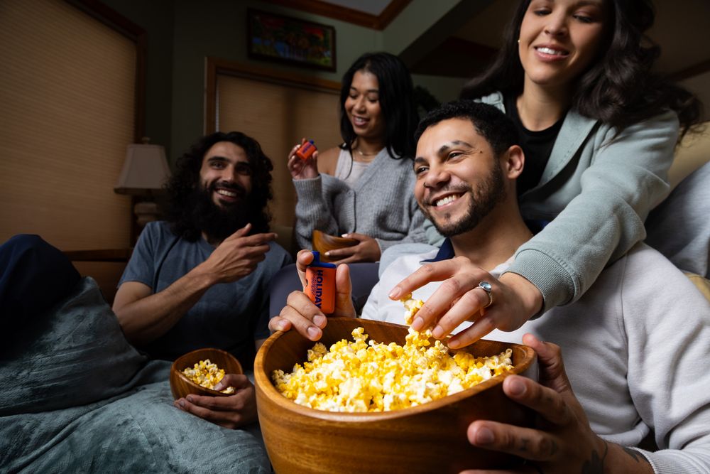 Friends laughing and sharing popcorn from a wooden bowl during a cozy movie night together indoors.