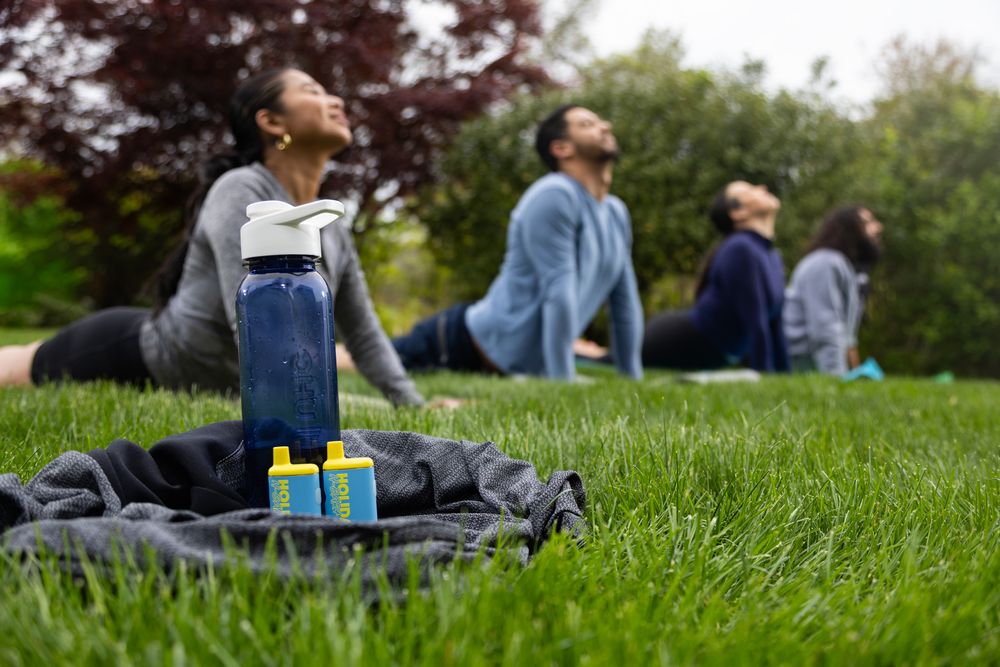 Group yoga class practicing cobra pose on grass in a park, with water bottles and a gym bag in the foreground.