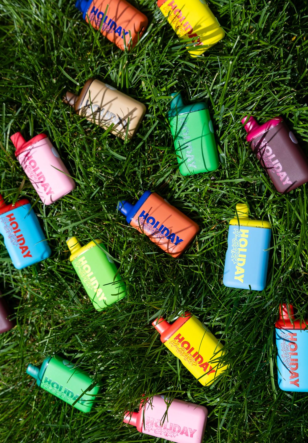 Colorful Holiday branded vape devices scattered on green grass in various bright colors including pink, blue, yellow, and green.
