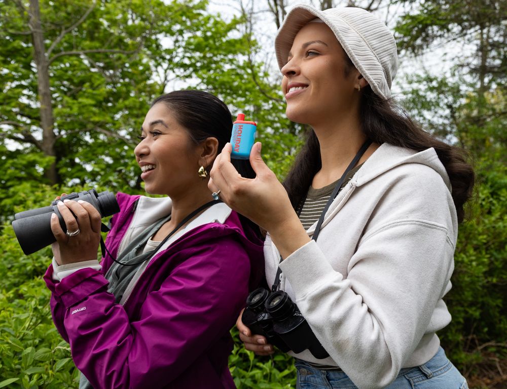 Two young women birdwatching outdoors, one holding binoculars and another with a water bottle, smiling in a lush green forest.