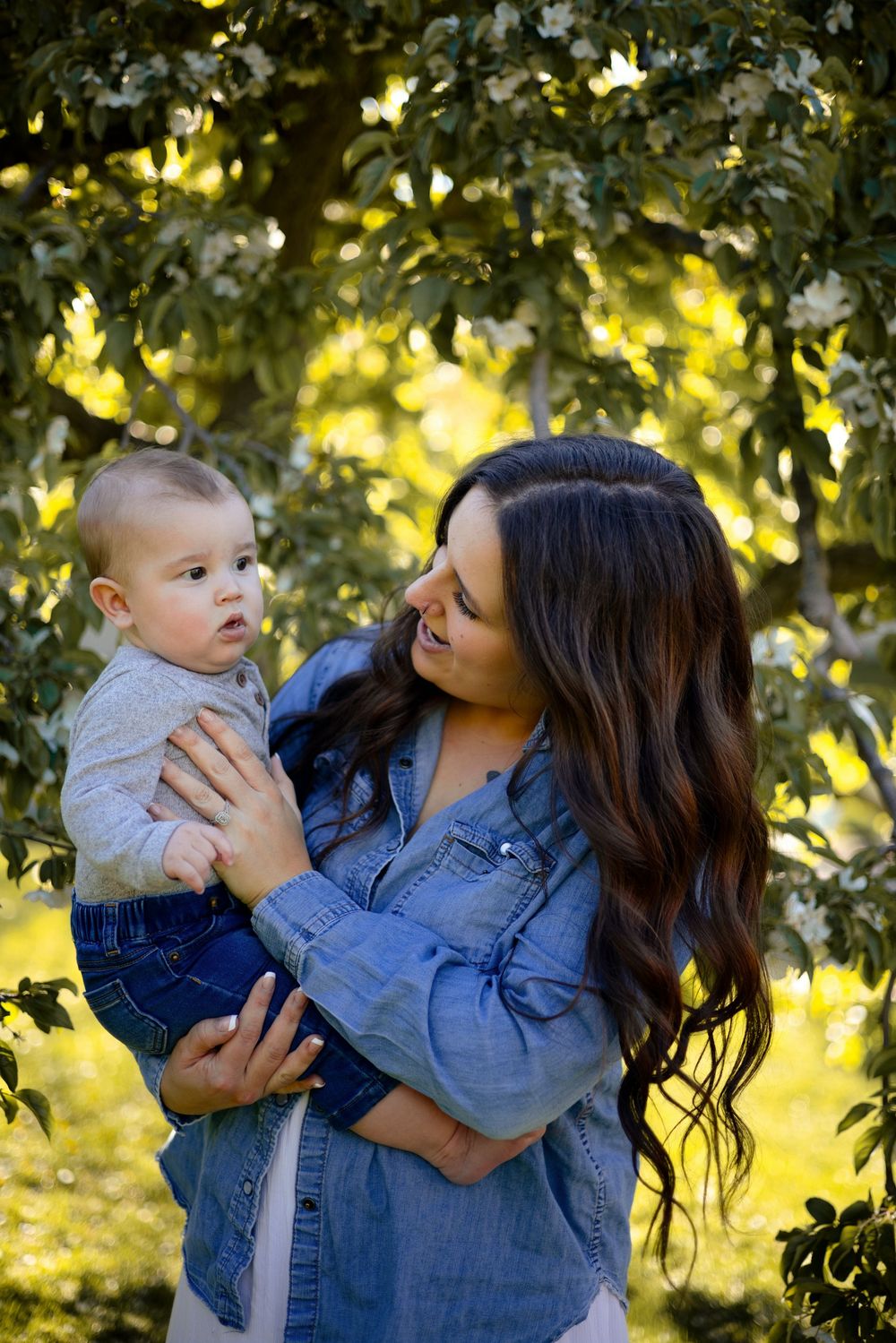 A young mother with long dark hair holds her baby boy outdoors beneath flowering trees in soft golden light.