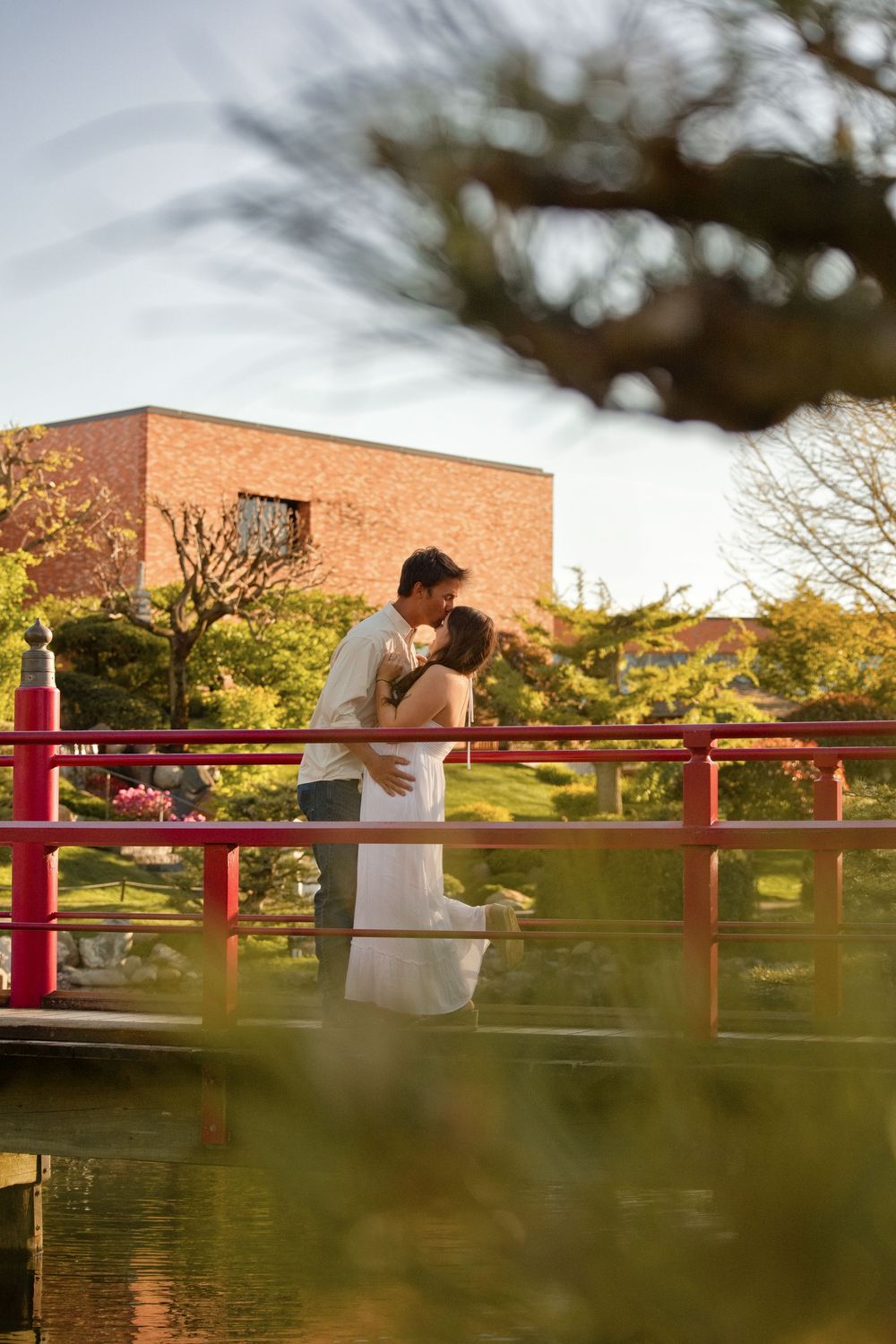 Couple sharing a romantic kiss on a red Japanese garden bridge surrounded by lush greenery and a brick building backdrop.