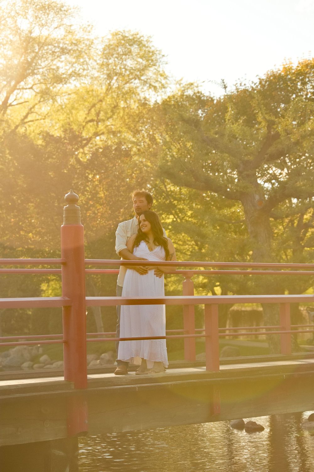 Couple in white standing on a red bridge at golden hour, surrounded by lush trees reflecting warm sunlight.