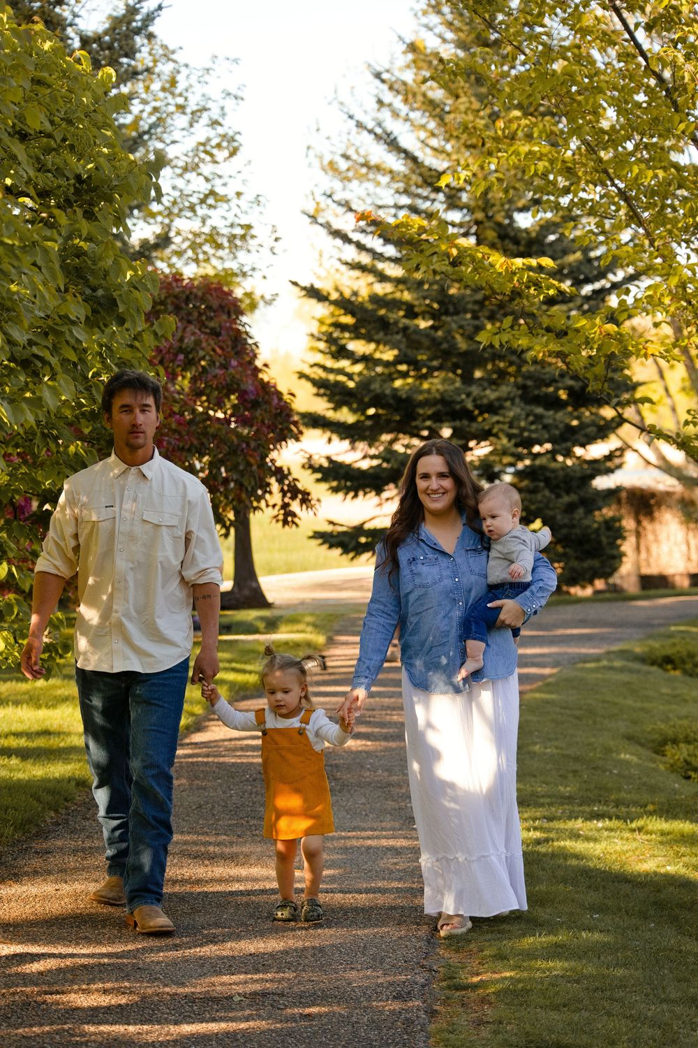 Family of four walking on a tree-lined path at golden hour, parents holding children's hands outdoors.