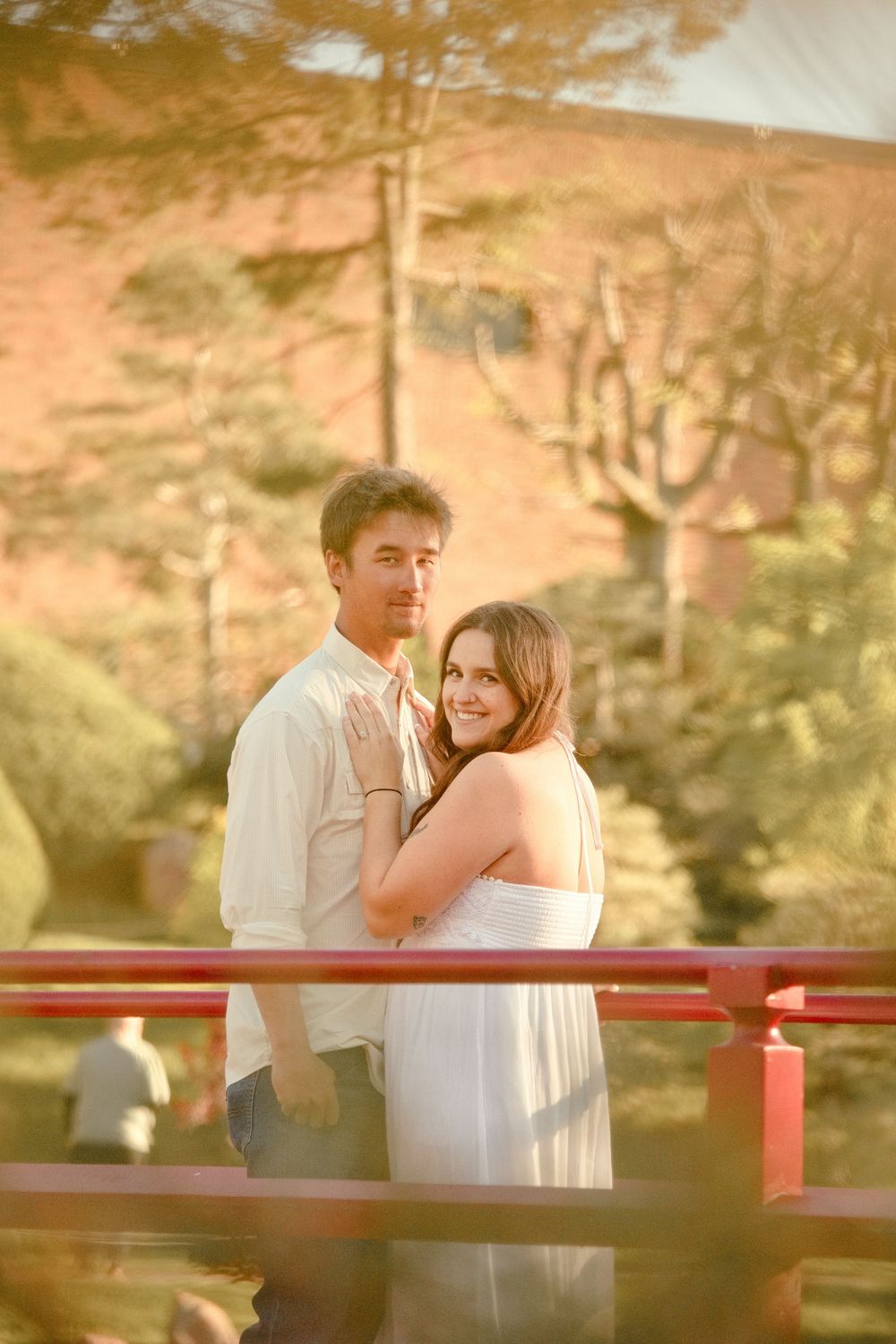 Couple embracing on a red bridge in a Japanese garden at golden hour, woman in white dress smiling.