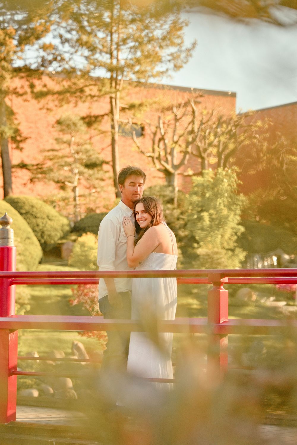 Couple embracing on a red bridge in a Japanese garden, bathed in warm golden sunlight with lush greenery.