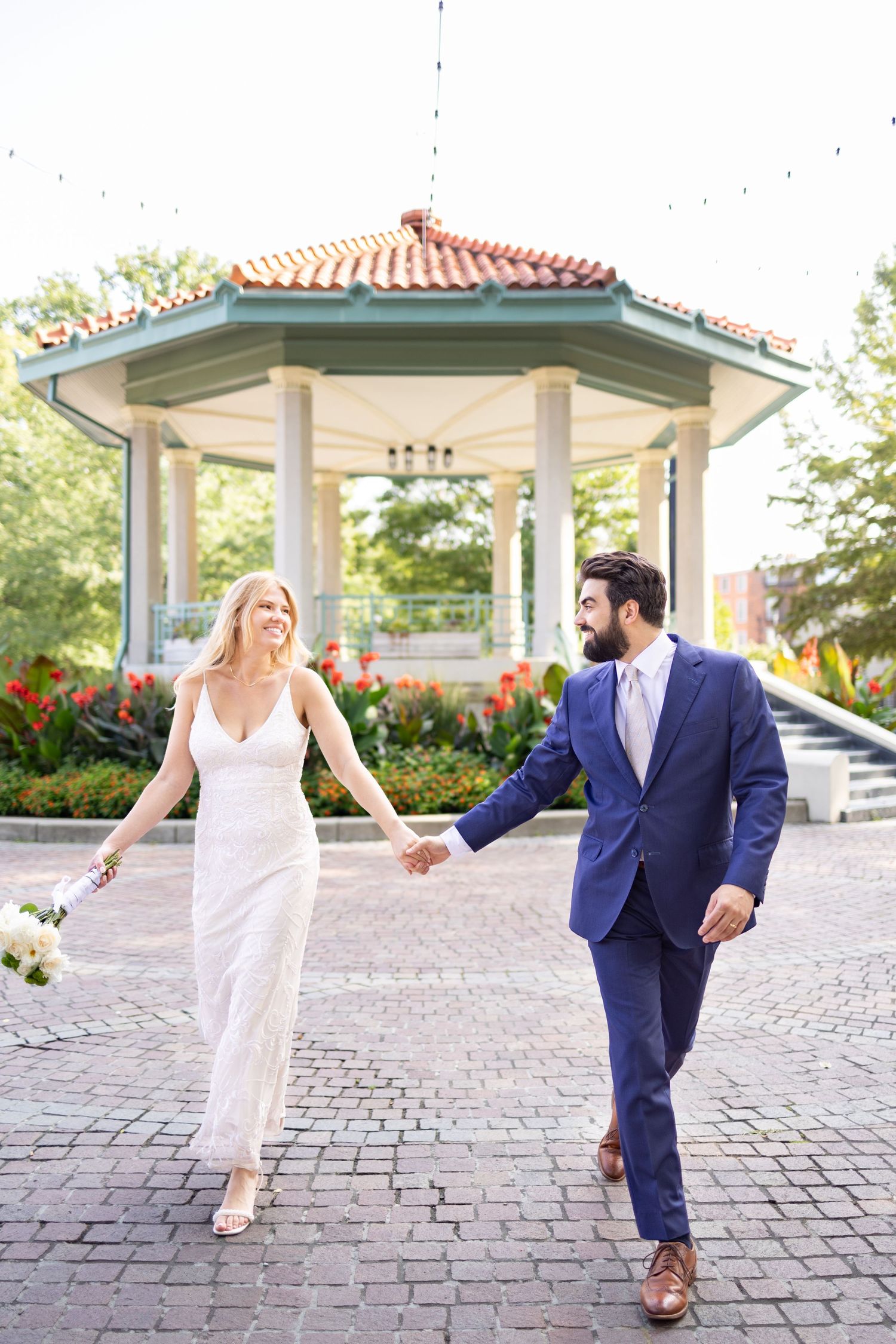 A smiling bride and groom hold hands while walking near a charming gazebo surrounded by lush greenery in Washington Park, Cincinnati Ohio.