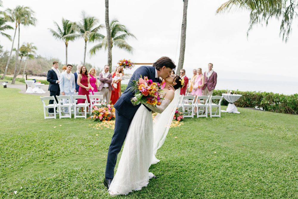 Bride and groom share a romantic dip kiss after their outdoor Hawaiian wedding ceremony with guests and palm trees behind them.