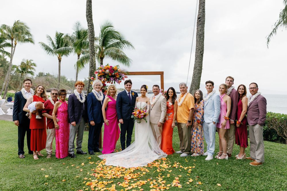 Wedding party group photo on tropical lawn with palm trees and floral arch.