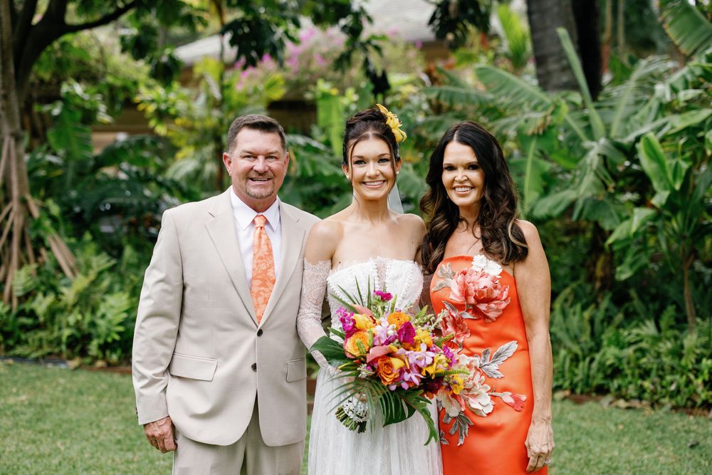 Bride holding tropical bouquet poses with parents in lush garden setting at Hawaiian wedding.