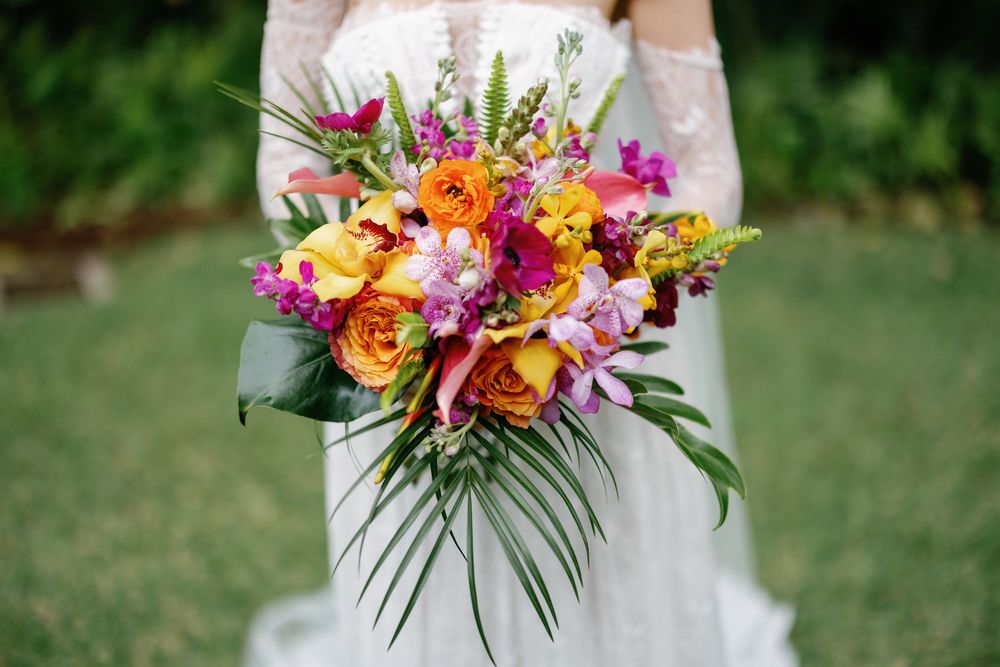 Bride holding a vibrant tropical wedding bouquet with orange, yellow, and pink flowers and lush green palm leaves.