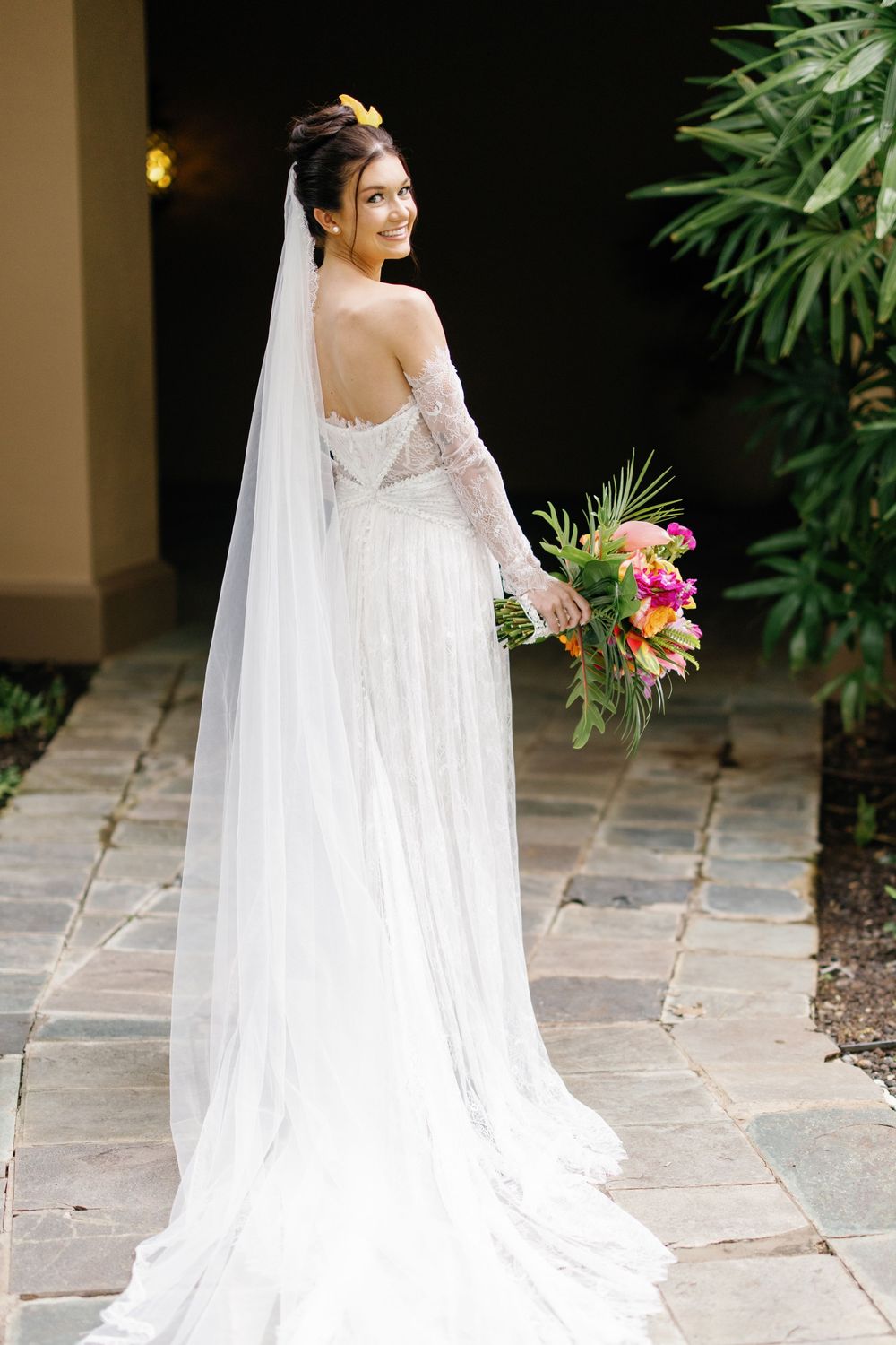 Bride in elegant lace wedding dress with long veil holding colorful tropical bouquet.