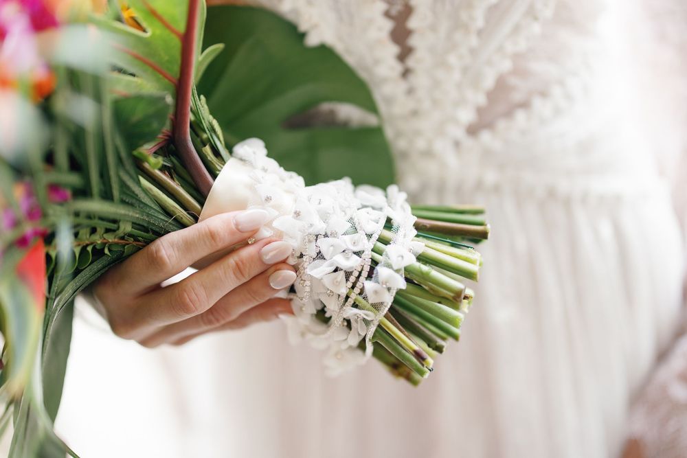 Bride holding a white floral bouquet with tropical green leaves, wearing a lace wedding dress with manicured nails.